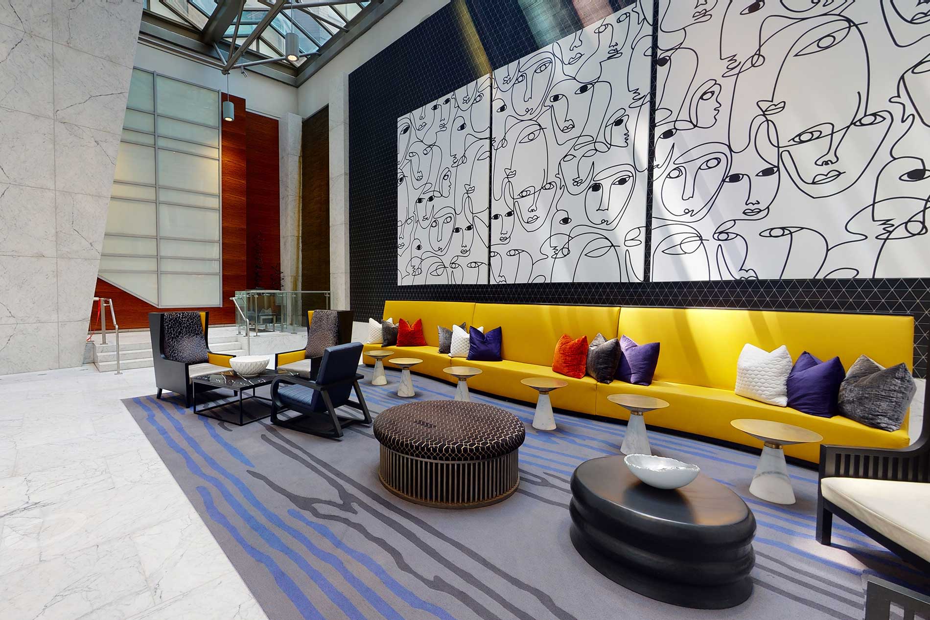 Modern lounge with bright yellow banquette seating, colorful throw pillows, geometric area rug, and abstract line-art wall panels beneath a skylight.