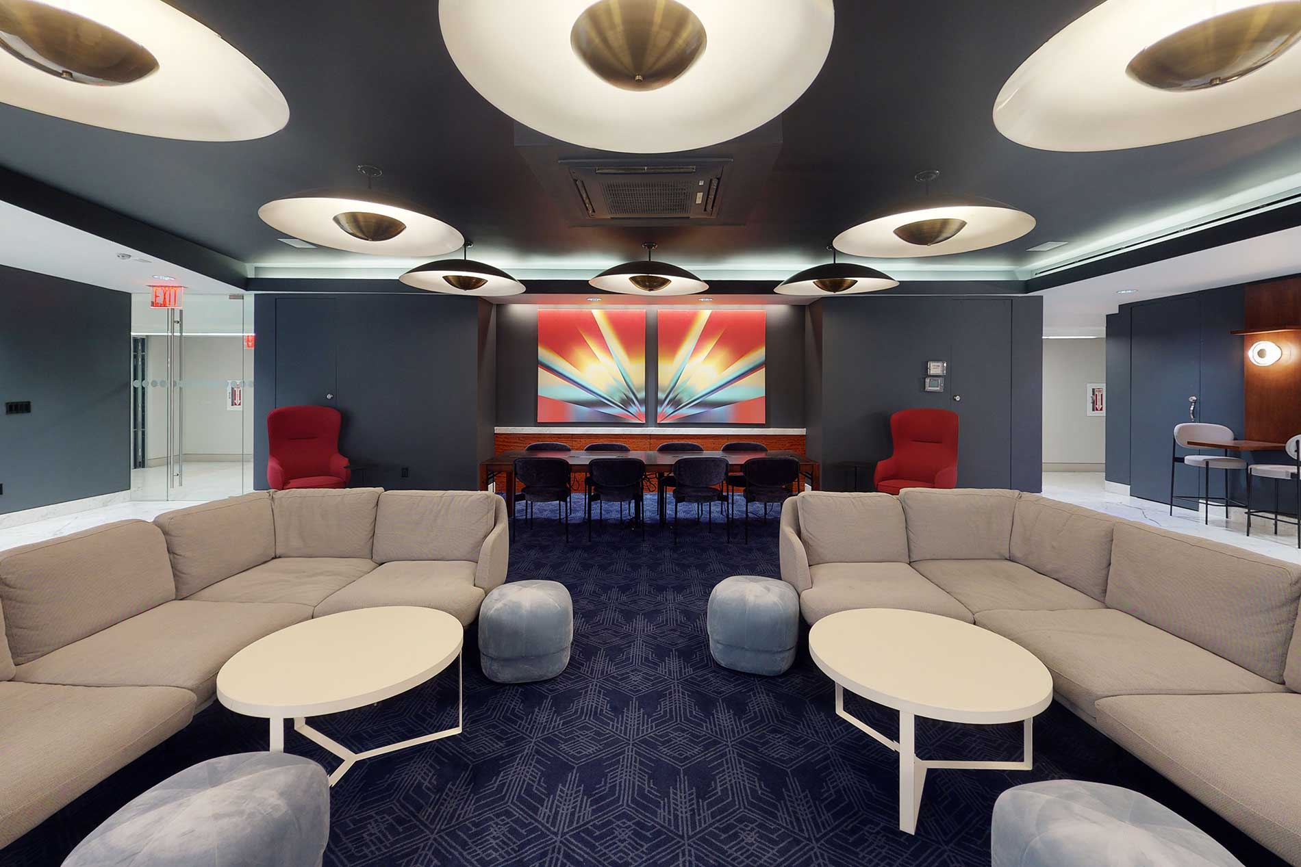 Modern lounge with two curved beige sectional sofas facing each other, white round tables, geometric ceiling lights, and colorful abstract artwork on dark blue walls.