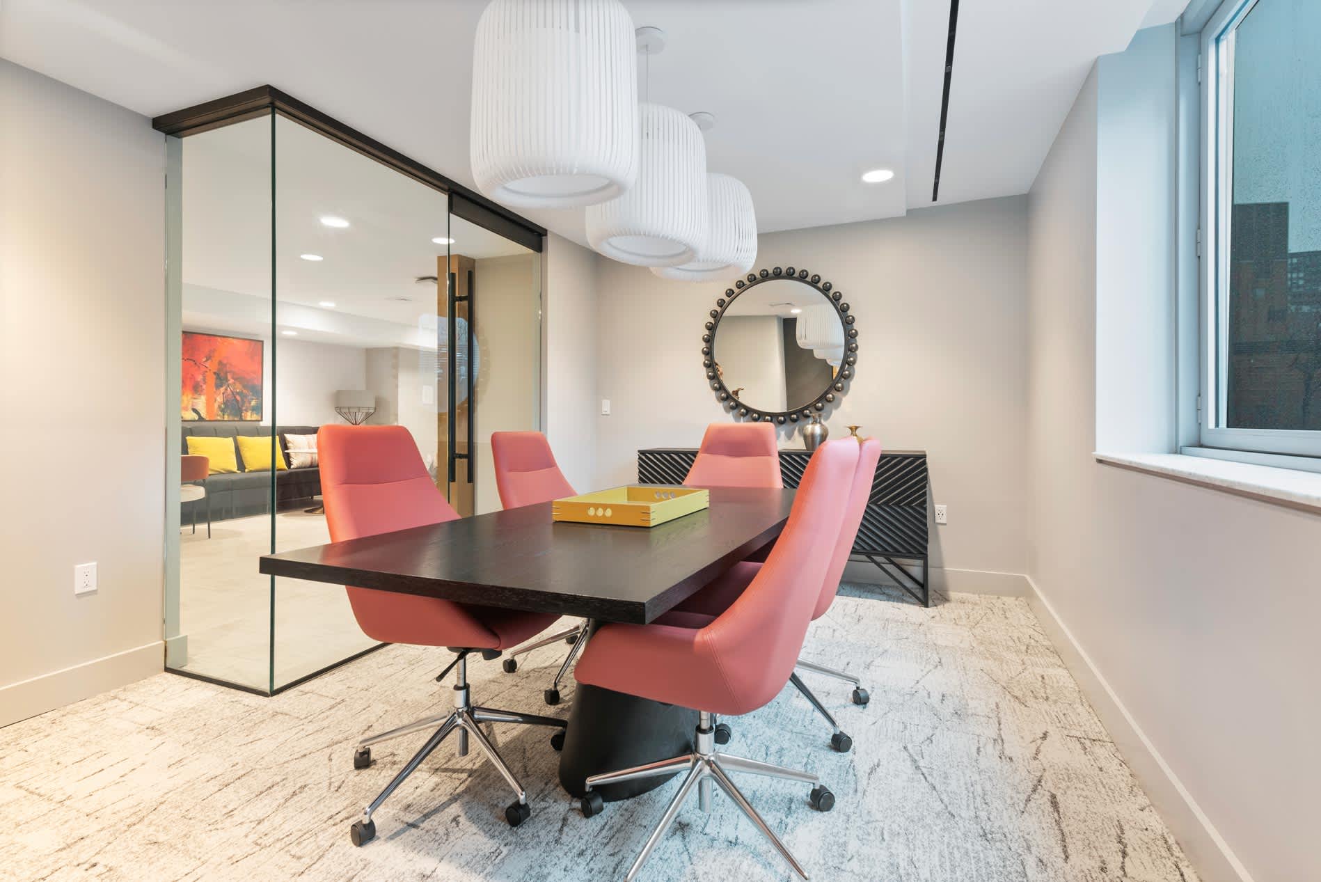 Conference room with black table, four coral office chairs, pendant lights, round wall mirror, and glass partition wall opening to adjacent space.
