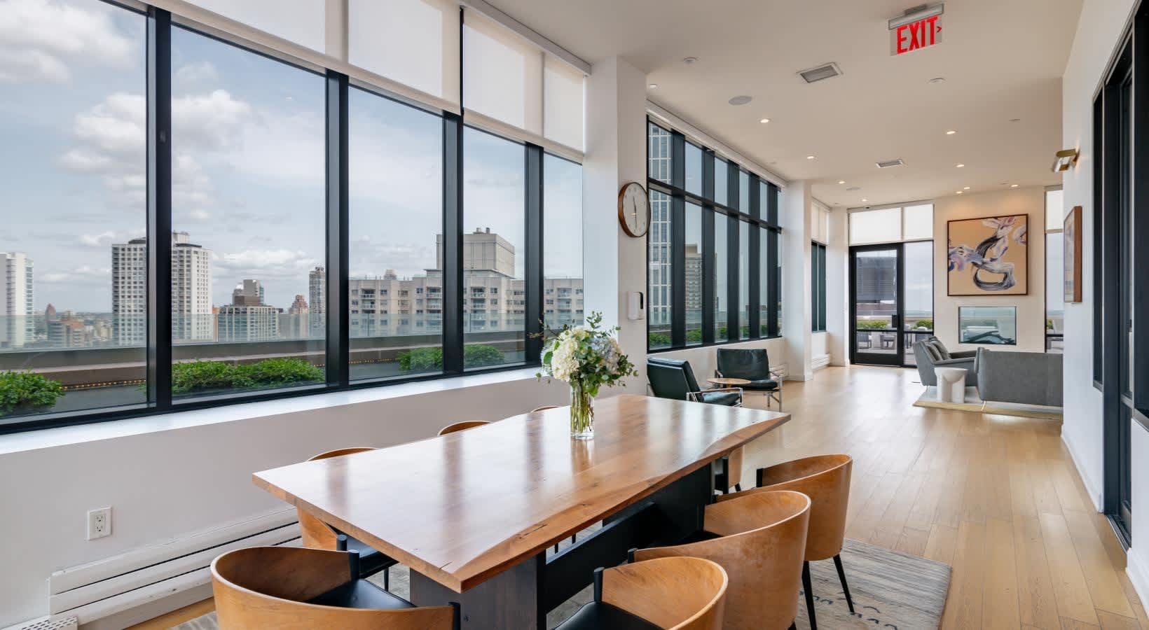 Common room with wood dining table, tan leather chairs, floor-to-ceiling windows with city views, light wood flooring, and modern furnishings.
