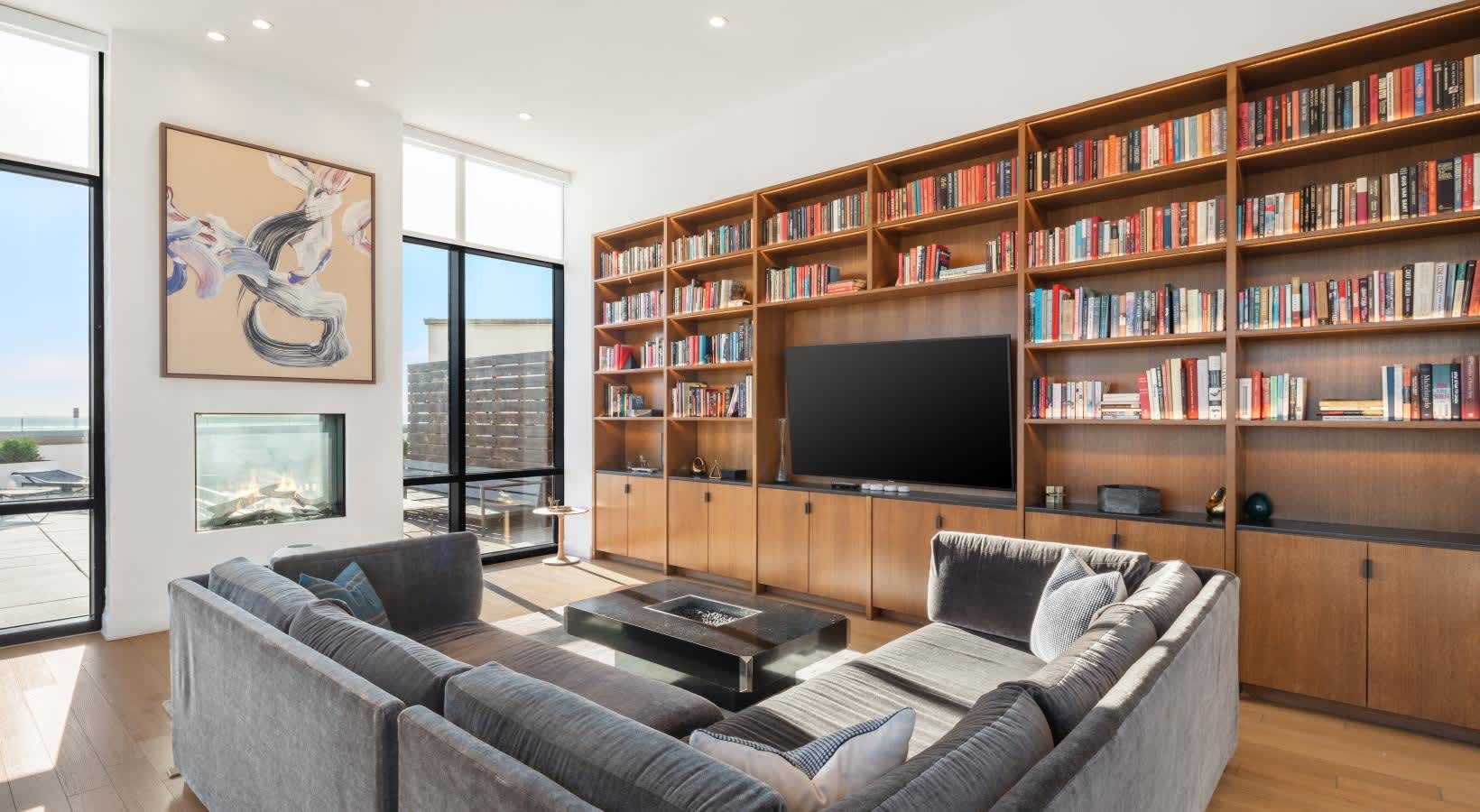 Living room with gray sectional sofa, built-in wood shelving with books and TV, large windows with water views, and hardwood flooring.