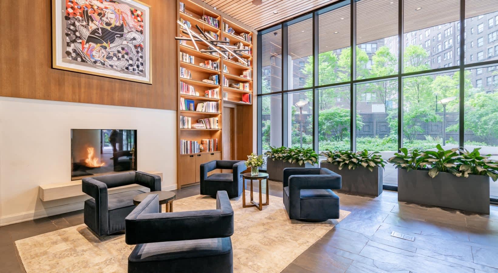 Modern living room with black curved seating, wooden shelving, framed artwork, potted plants, and expansive grid windows overlooking urban buildings.