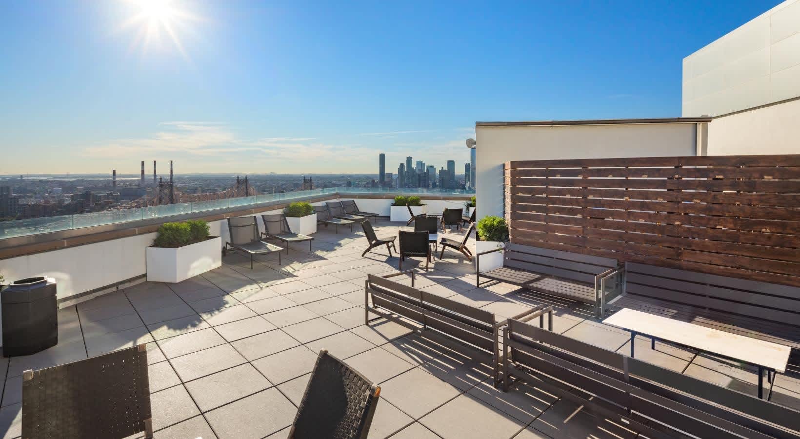 Rooftop terrace with lounge chairs, benches, planters, and wooden privacy screen overlooking New York City skyline.