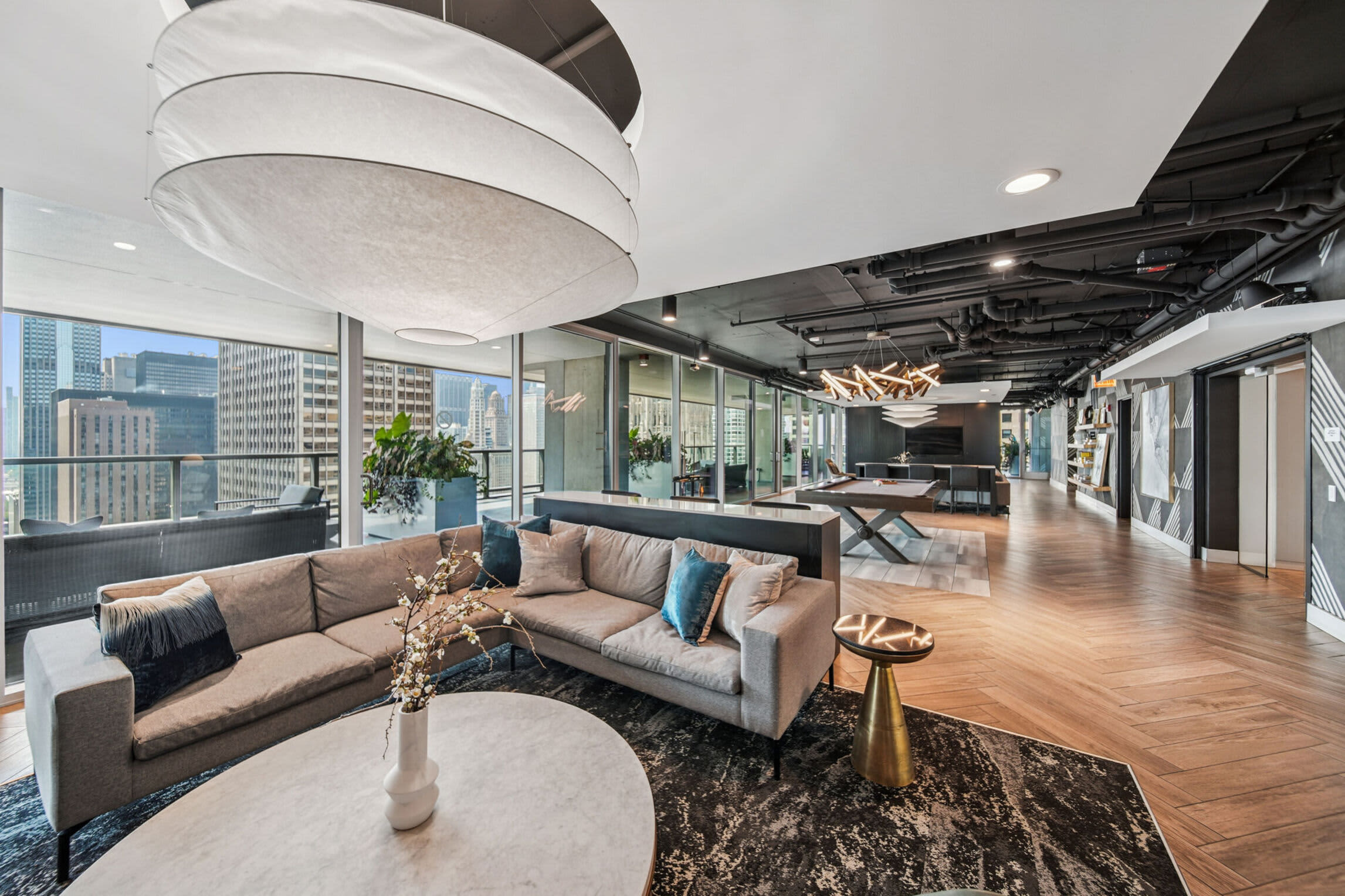 High-rise living room with taupe L-shaped sofa, round marble coffee table, floor-to-ceiling windows showing Chicago skyline, hardwood floors, and modern pendant lighting.