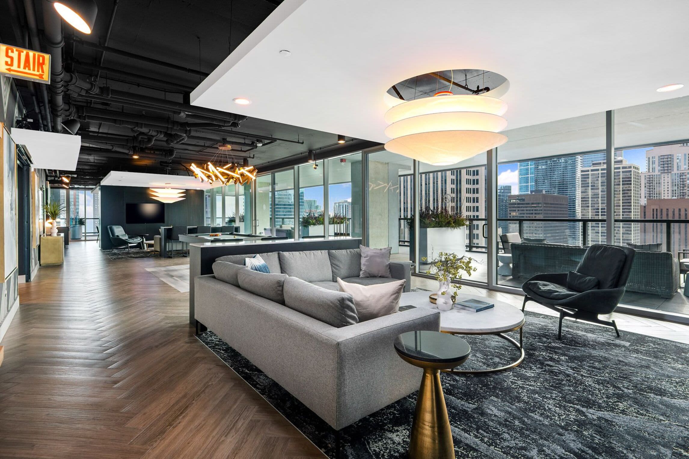 Open-concept living room with gray sectional sofa, modern ceiling fixture, hardwood floors, and expansive windows showing Chicago skyline views.