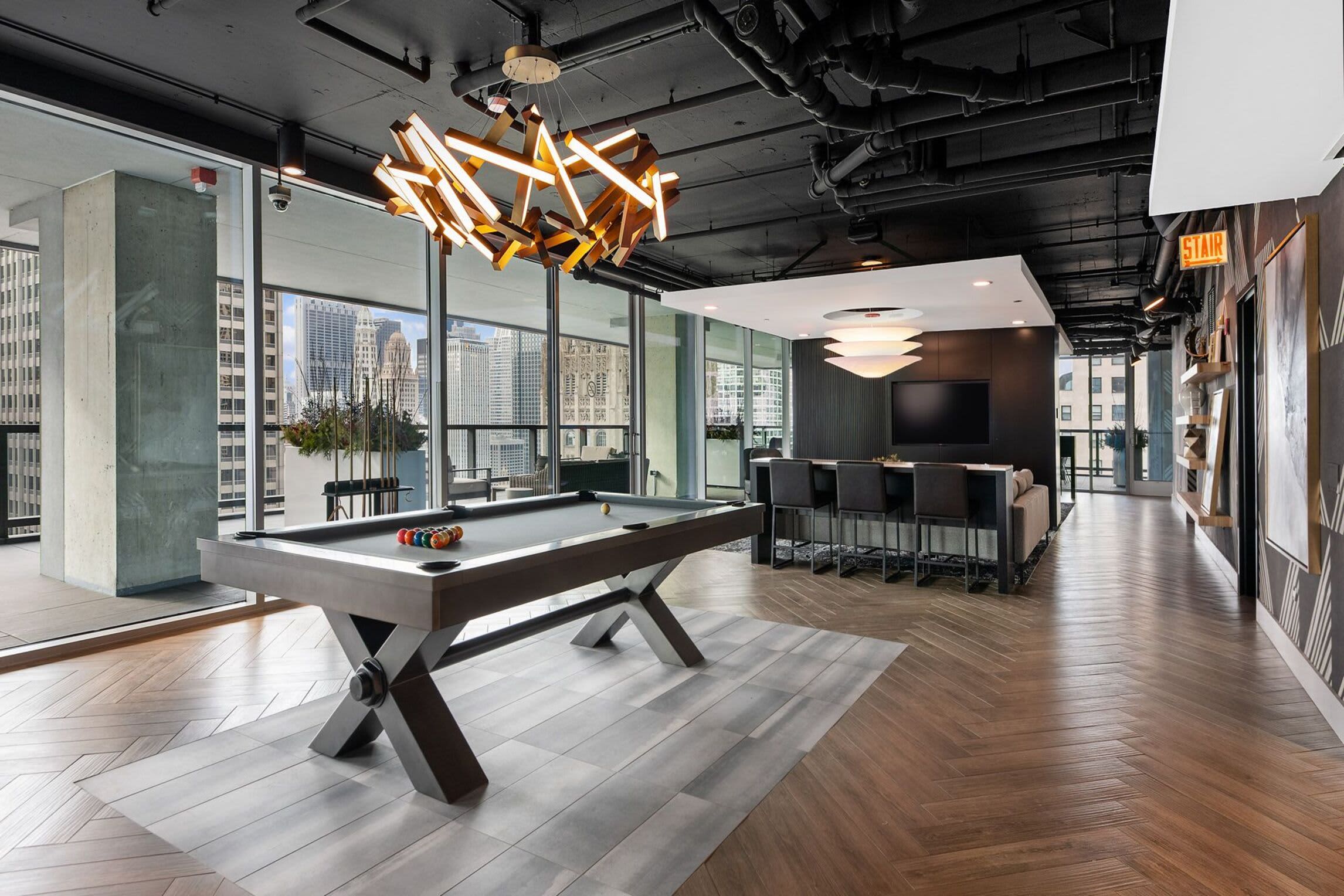 Recreation room with pool table, modern gold pendant light fixture, wood and tile flooring, and floor-to-ceiling windows overlooking Chicago skyline.