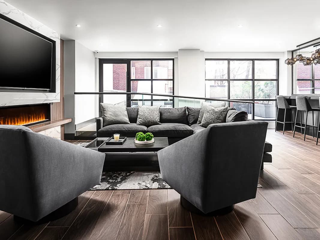 Contemporary living room with gray upholstered chairs, dark sofa, electric fireplace with TV above, wood flooring, and large windows overlooking brick buildings.