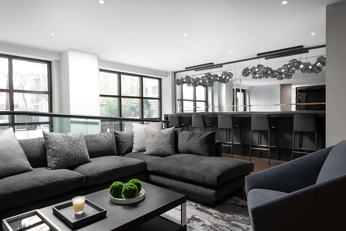 Modern living room with dark gray sectional sofa, black coffee table, kitchen island with bar seating, and large windows with city views.