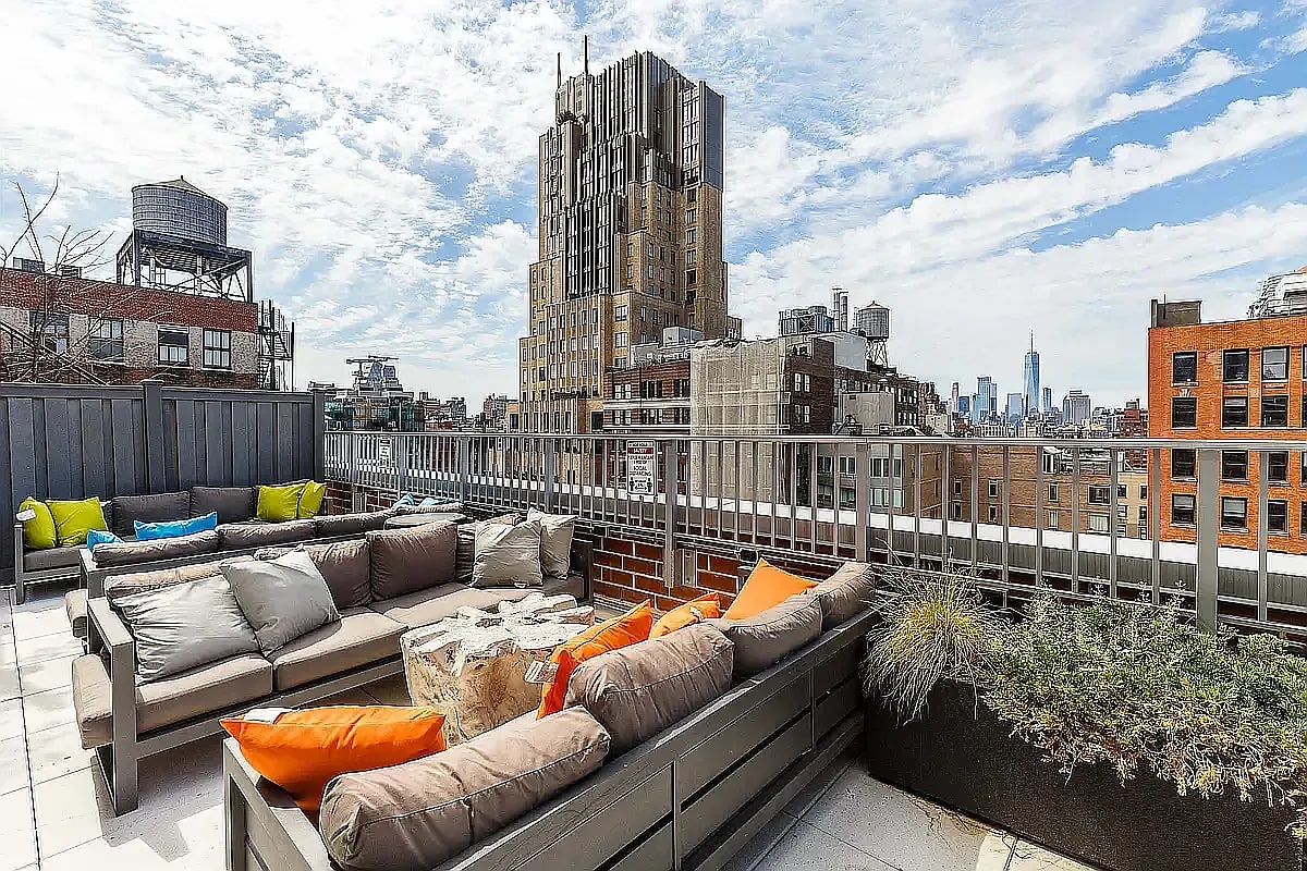 Rooftop terrace with gray lounge seating, orange pillows, potted plants, and white metal railing overlooking Manhattan skyline.