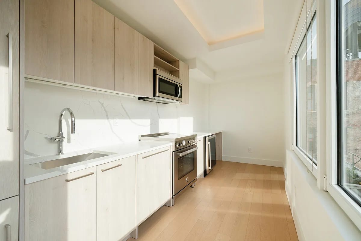 Galley kitchen with light wood cabinets, white marble backsplash, stainless steel oven and sink, and large windows with natural light.