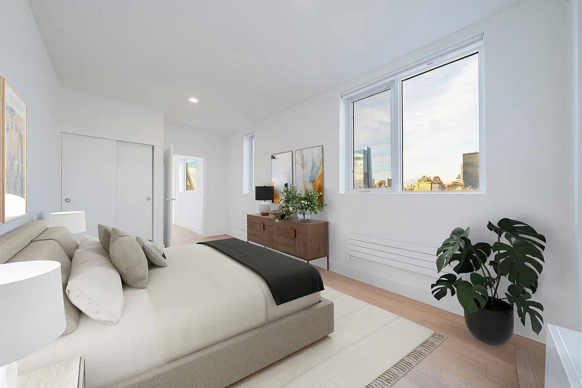Modern bedroom with cream upholstered bed, large windows with city views, wood dresser, potted plant, and light wood flooring.
