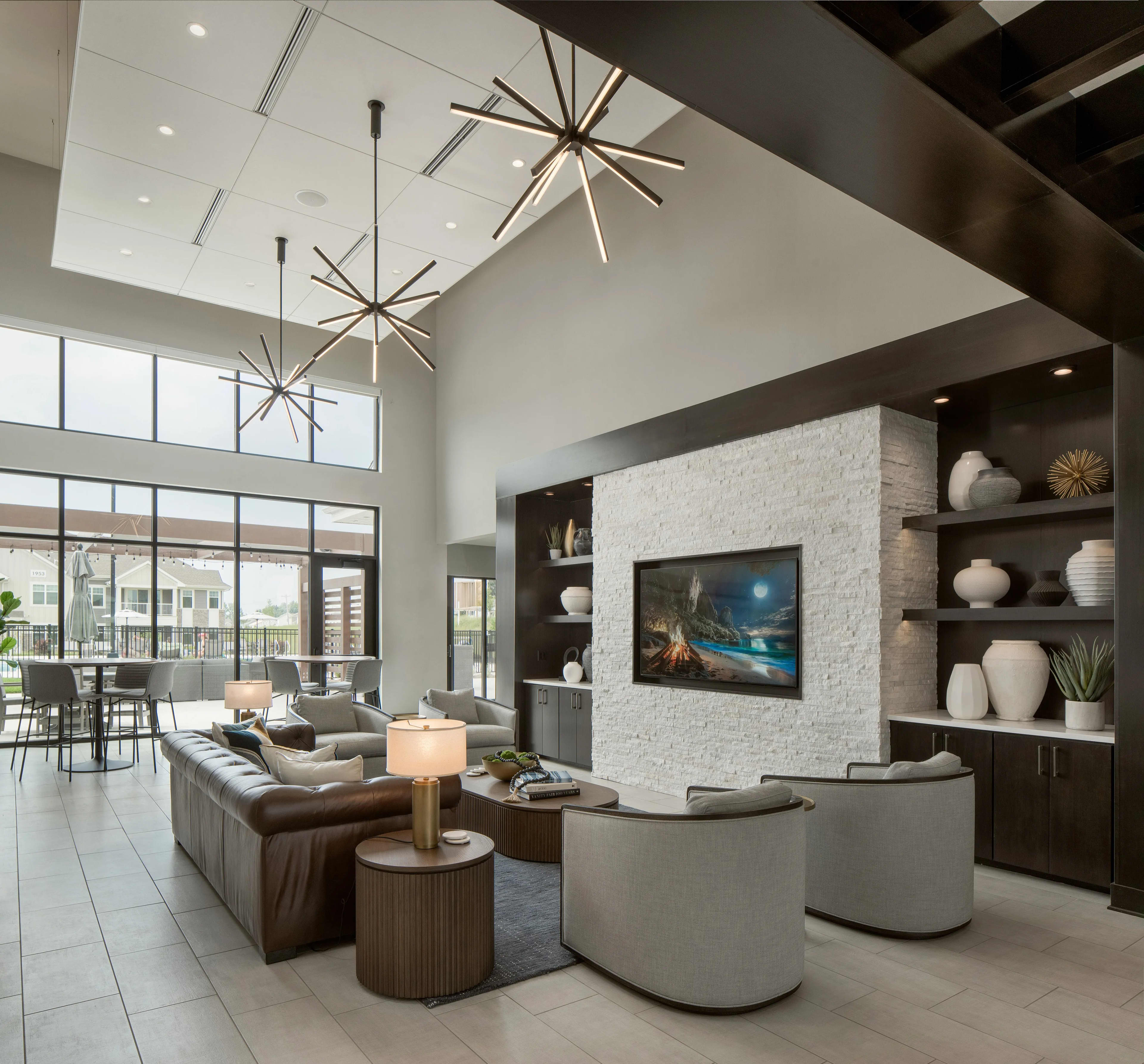 Modern lounge with brown leather sofa, white brick fireplace with TV, dark shelving, geometric pendant lights, and floor-to-ceiling windows overlooking a balcony.