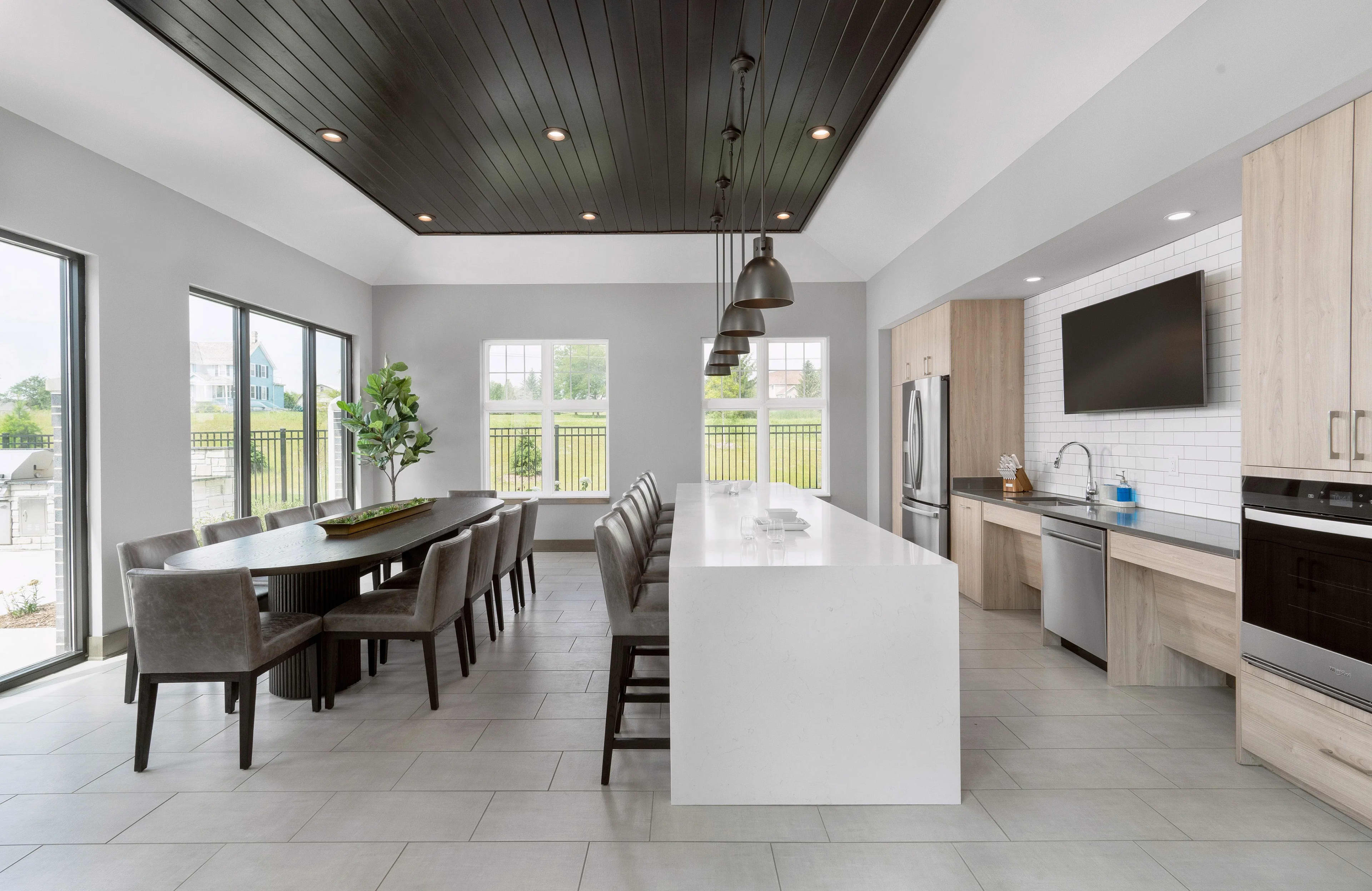 Modern kitchen and dining room with white island, gray dining chairs, stainless steel appliances, dark wood ceiling, and multiple windows overlooking green landscape.