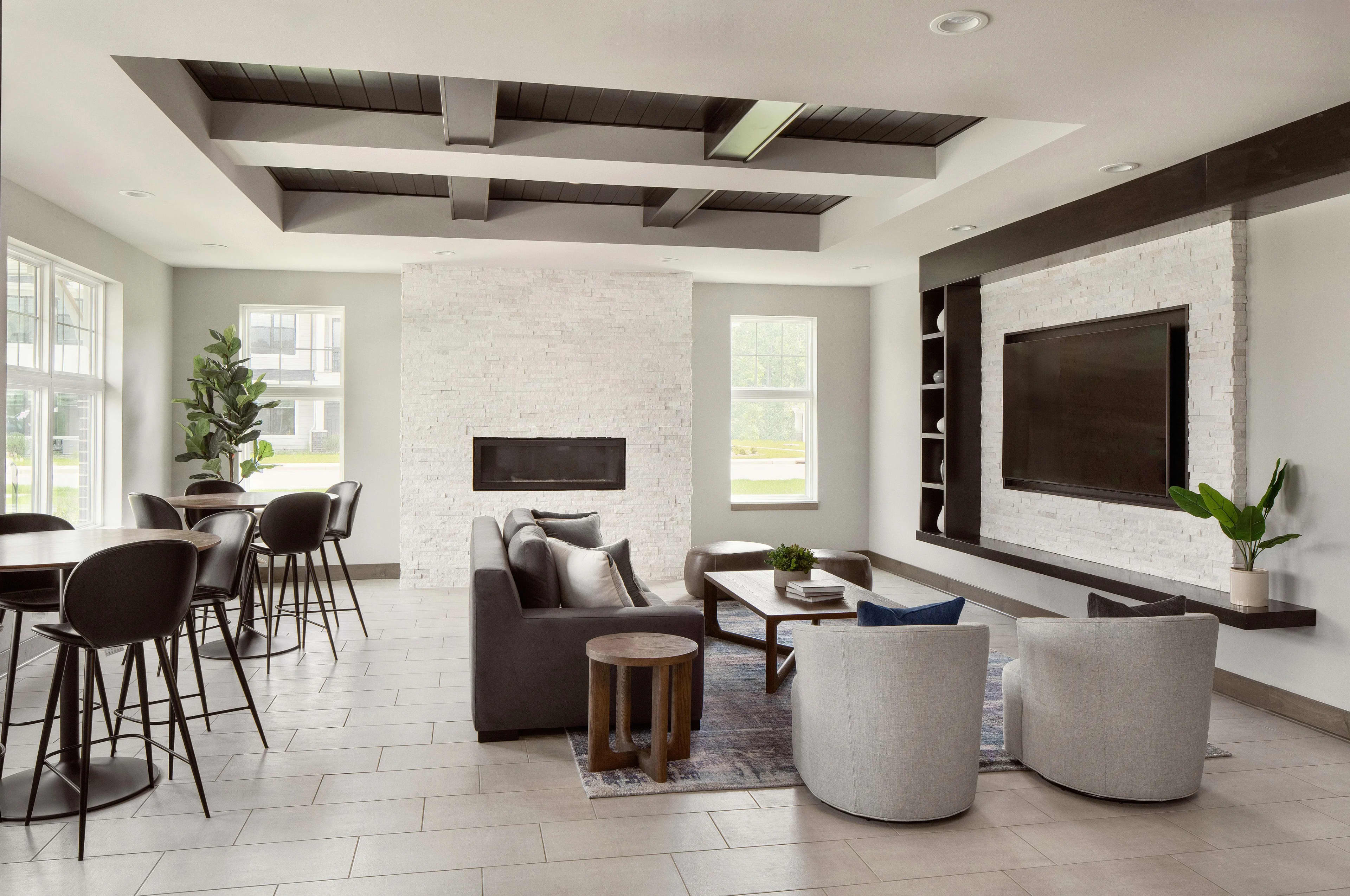 Open-concept living room with white brick fireplace, wall-mounted TV, coffered ceiling, bar seating, and contemporary furniture in neutral tones.