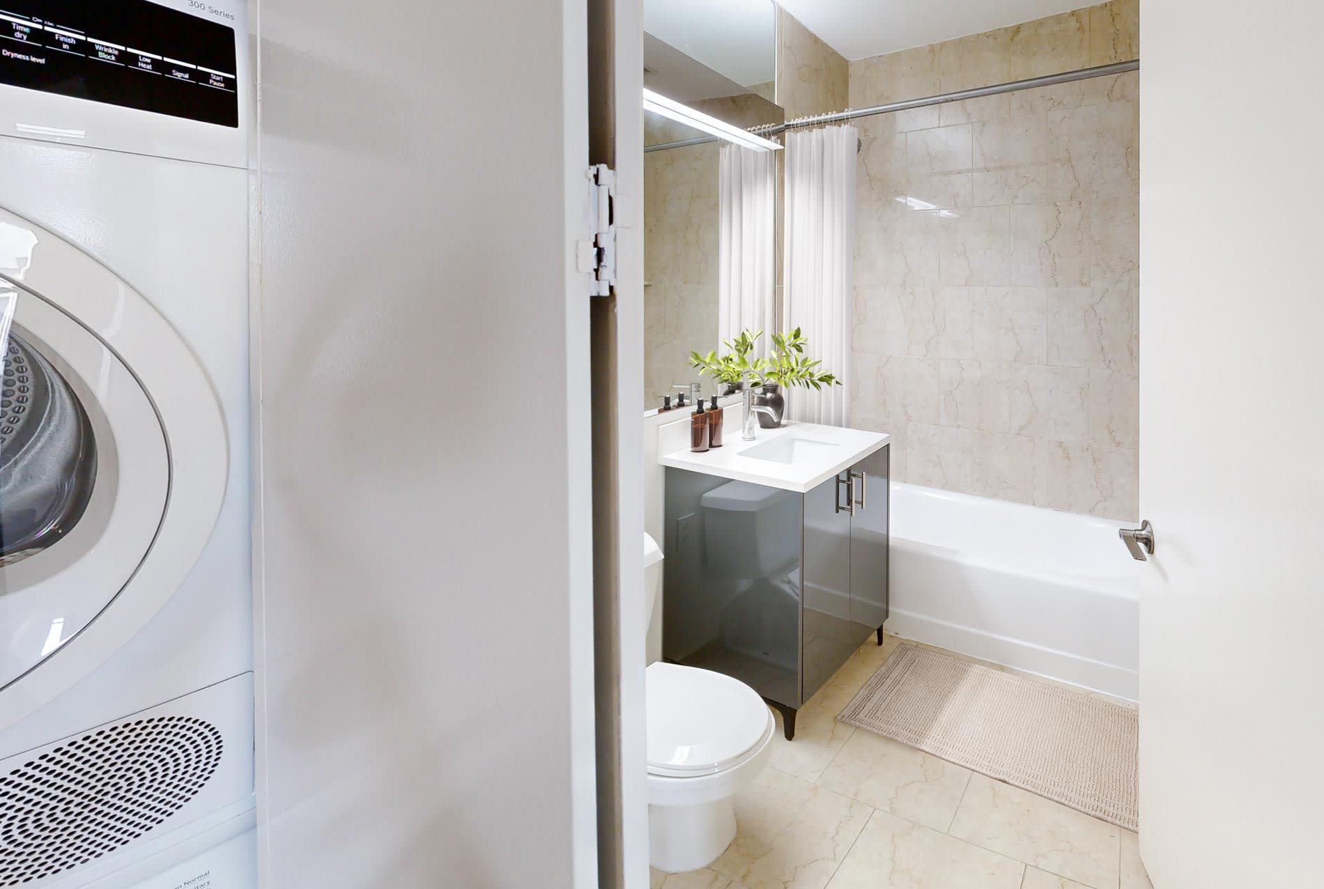 Bathroom with marble tile walls, white soaking tub, dark vanity with white sink, mirror, potted plant, and white washer-dryer visible on left.