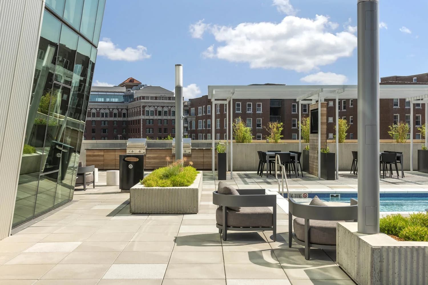 Rooftop terrace with gray lounge chairs, planters, dining tables, pool, and downtown brick buildings in background.