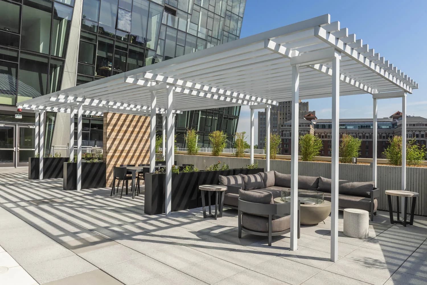 Rooftop terrace with white pergolas, gray lounge seating, planters, and city skyline views.