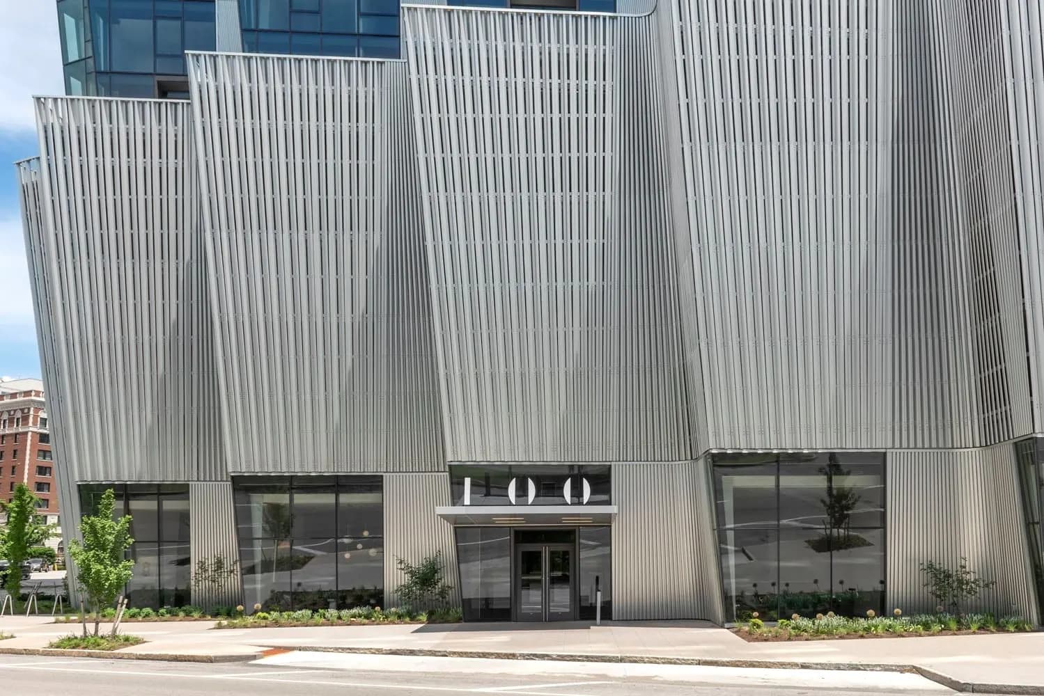 Modern building exterior with vertical corrugated metal facade, large ground-floor windows, and central entrance marked with 100 address numbers.