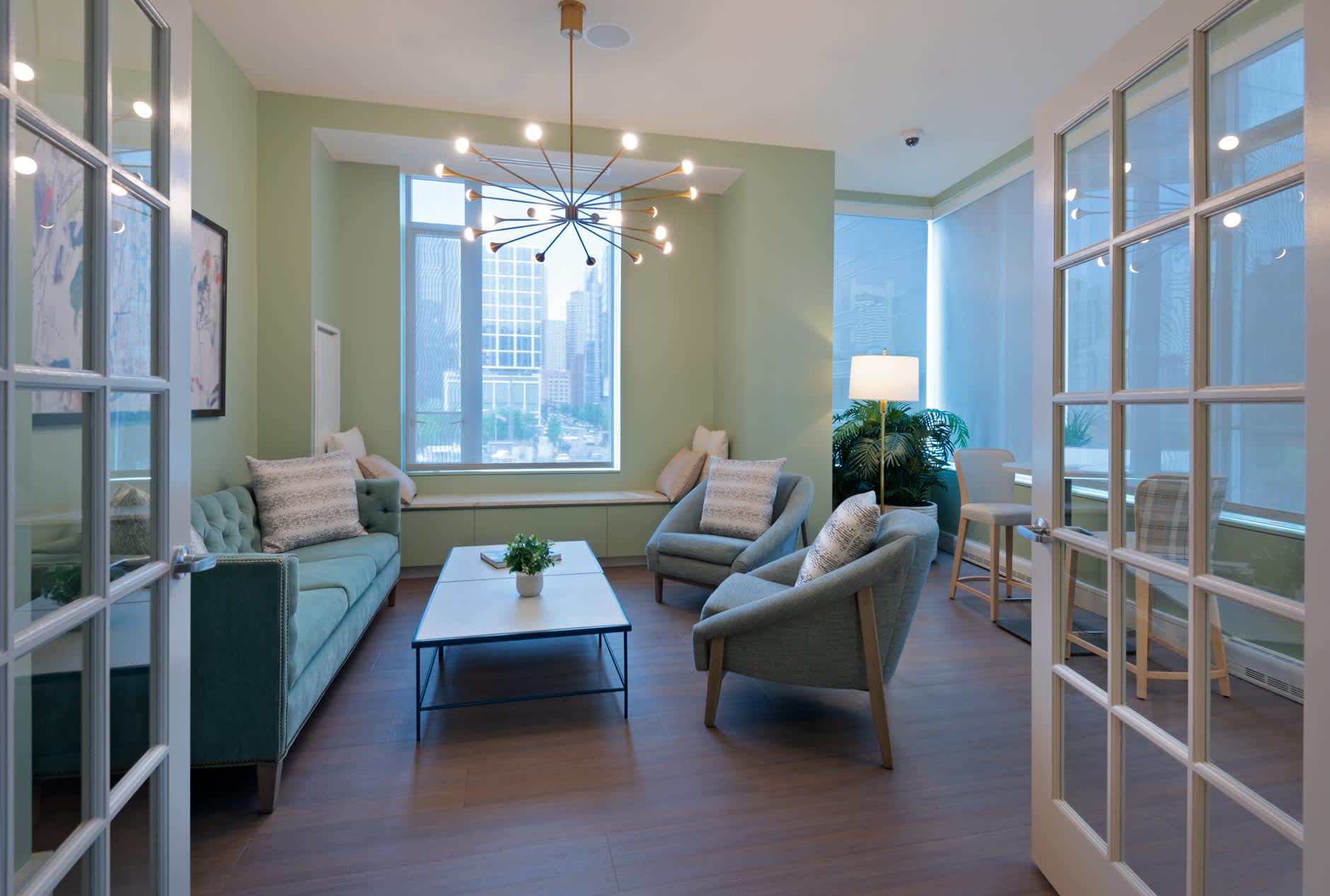 Modern lounge with sage green accent wall, blue-gray seating, geometric pendant light, and expansive windows overlooking downtown Boston skyline.