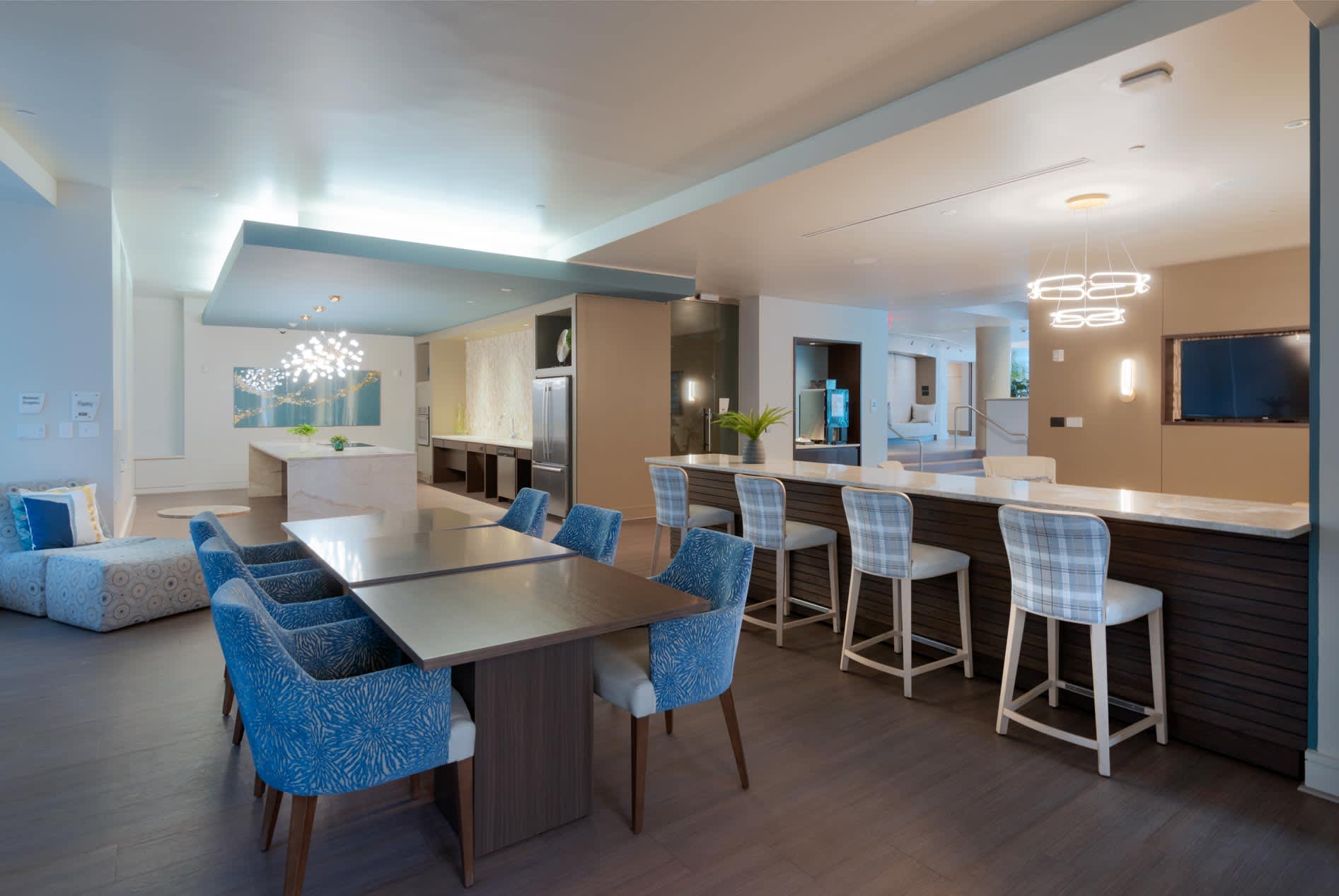 Open common area with dining table surrounded by blue patterned chairs, bar seating, kitchen, and living space with contemporary lighting.