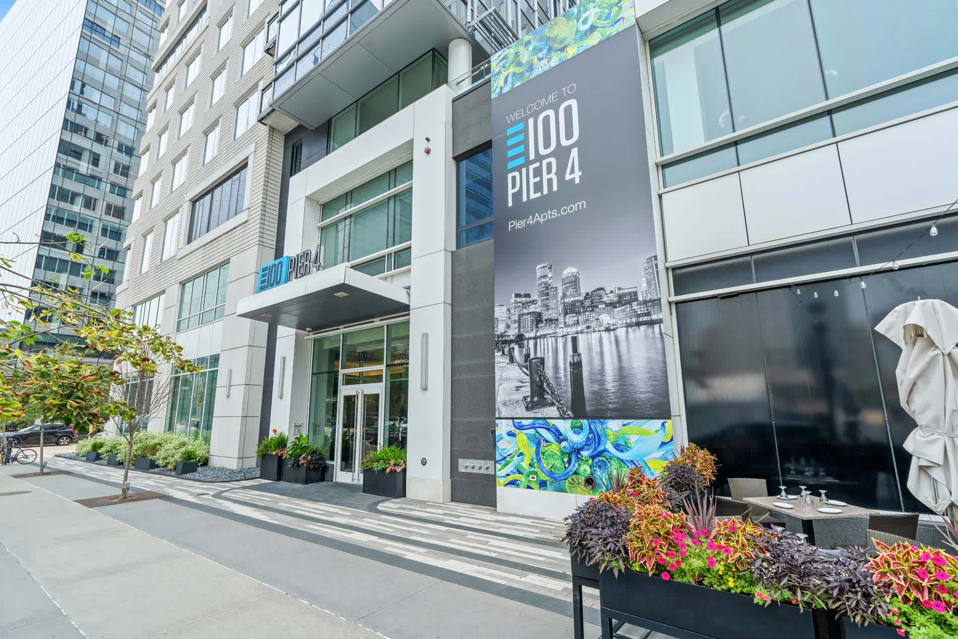 Modern residential building entrance with glass doors, white facade, colorful flower planters, and welcome signage for 100 Pier 4.