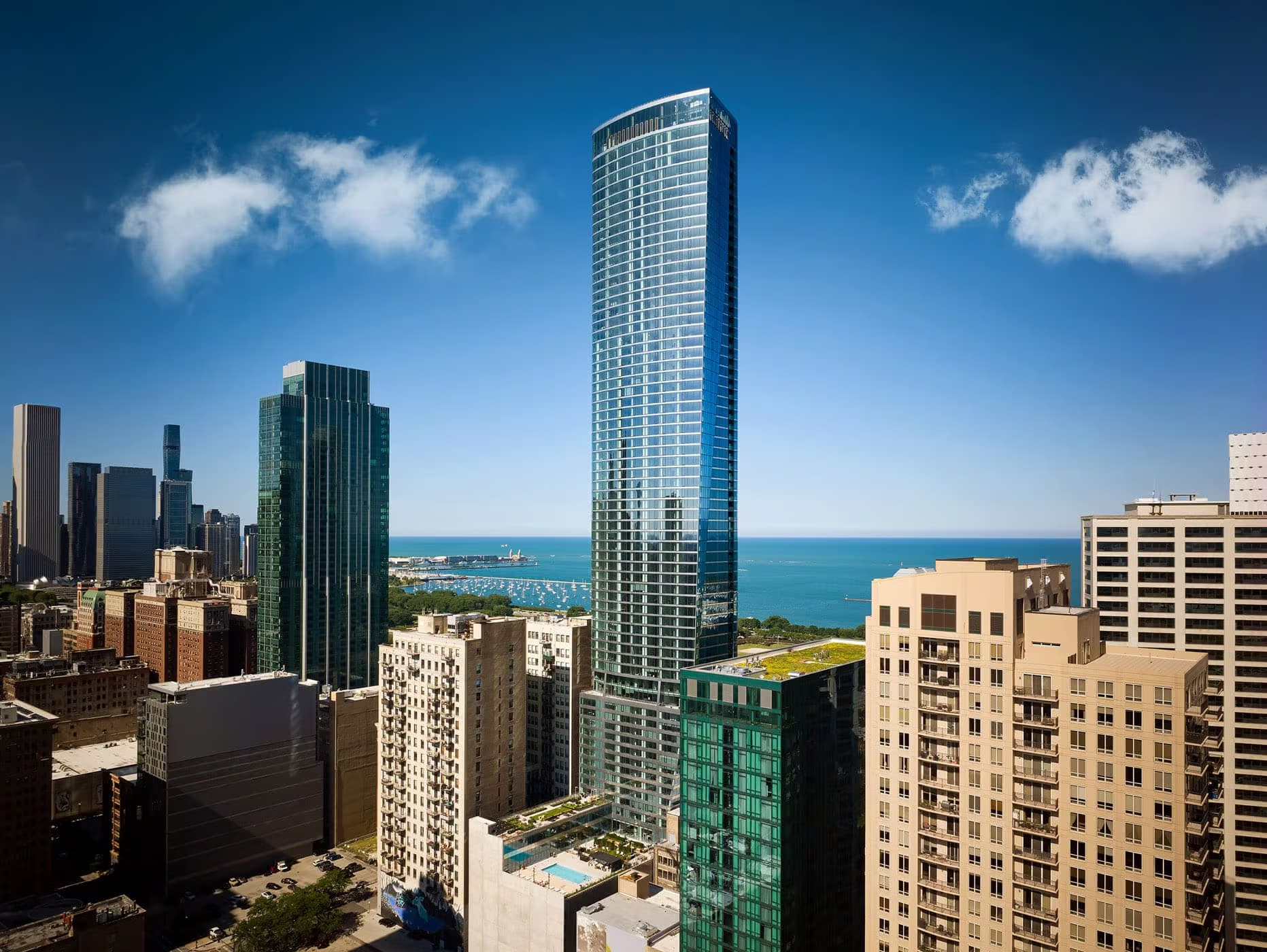 Tall residential tower with floor-to-ceiling windows overlooking Lake Michigan, surrounded by Chicago skyline buildings under blue sky.