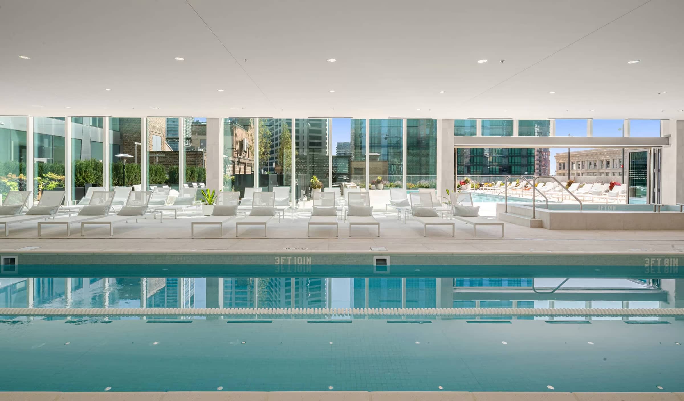 Indoor lap pool with turquoise water, white lounge chairs along the deck, floor-to-ceiling windows, and city views.
