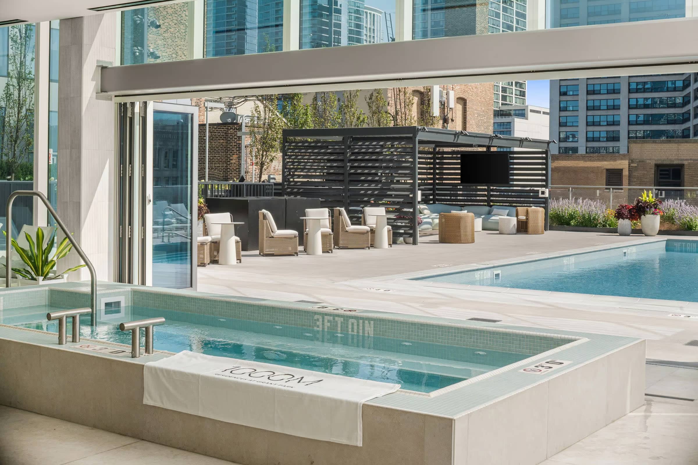 Indoor soaking tub with glass doors opening to rooftop pool, lounge chairs, and Chicago skyline views.