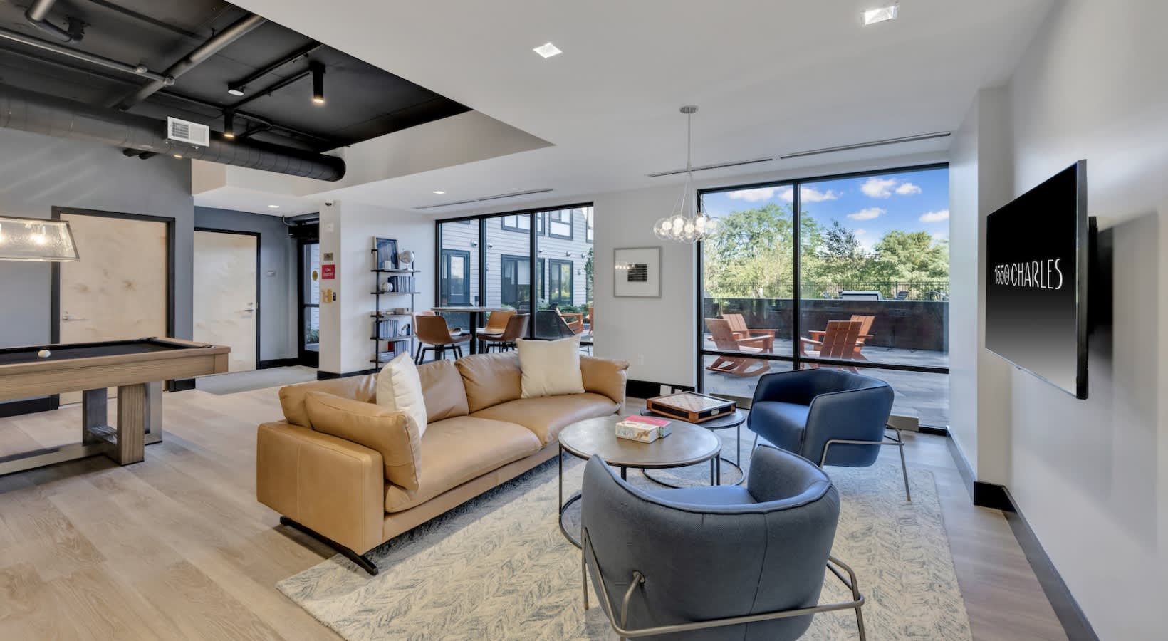 Modern lounge with tan sofa, blue accent chairs, glass doors opening to rooftop terrace with green views, and exposed ceiling beams.