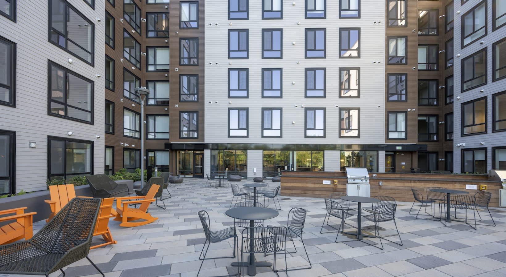 Outdoor courtyard with orange and black lounge chairs, dining tables, built-in grill, and paved patio surrounded by multi-story residential buildings.