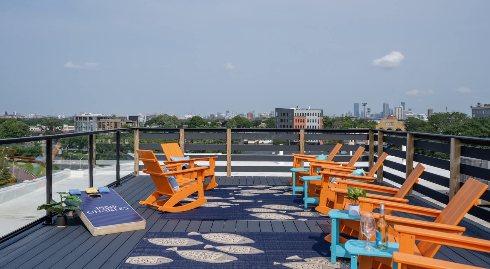 Rooftop deck with orange Adirondack chairs, blue accents, cornhole game, and Boston skyline view across the Charles River.
