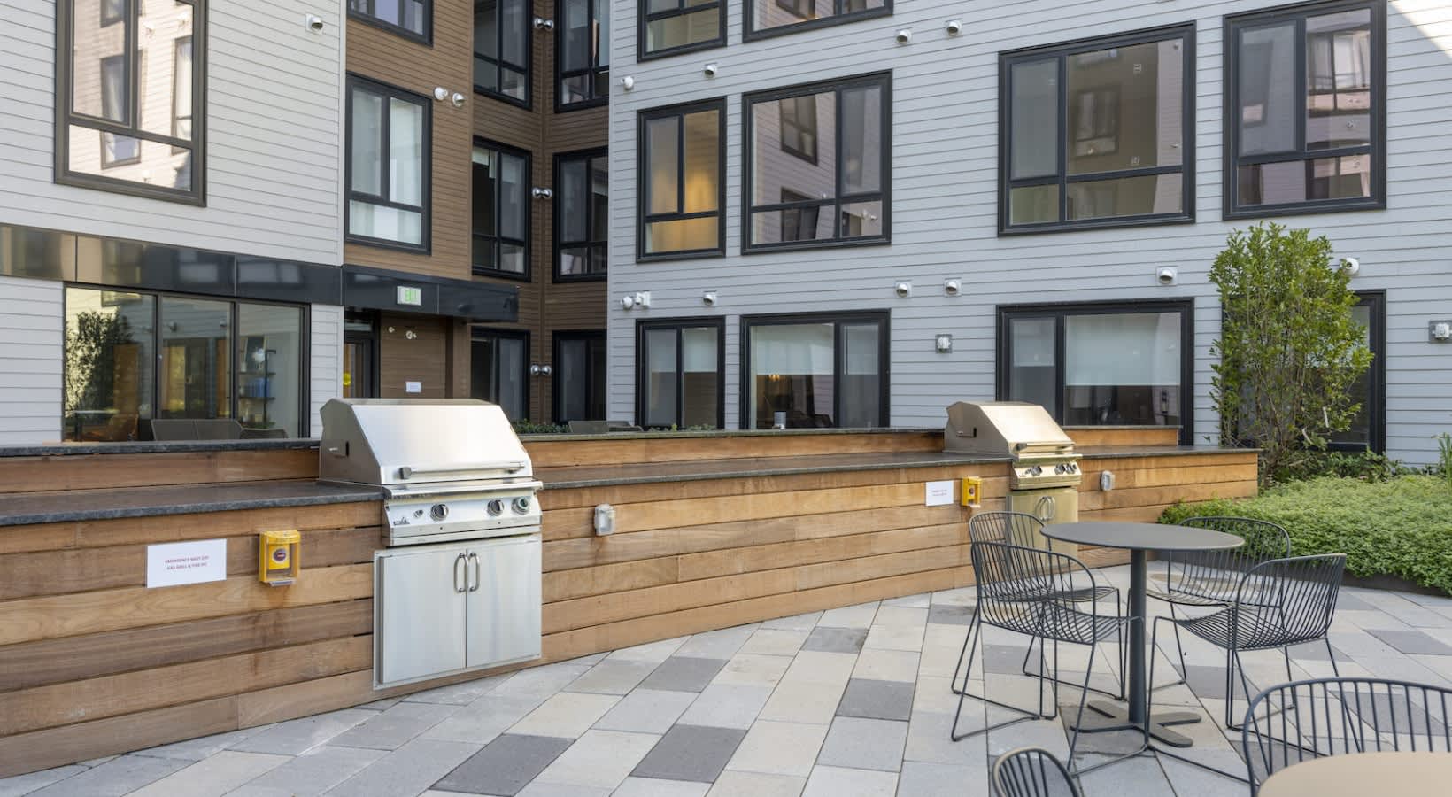 Outdoor courtyard with stainless steel grills, wooden counter, black dining chairs, and multi-story residential building with white siding.