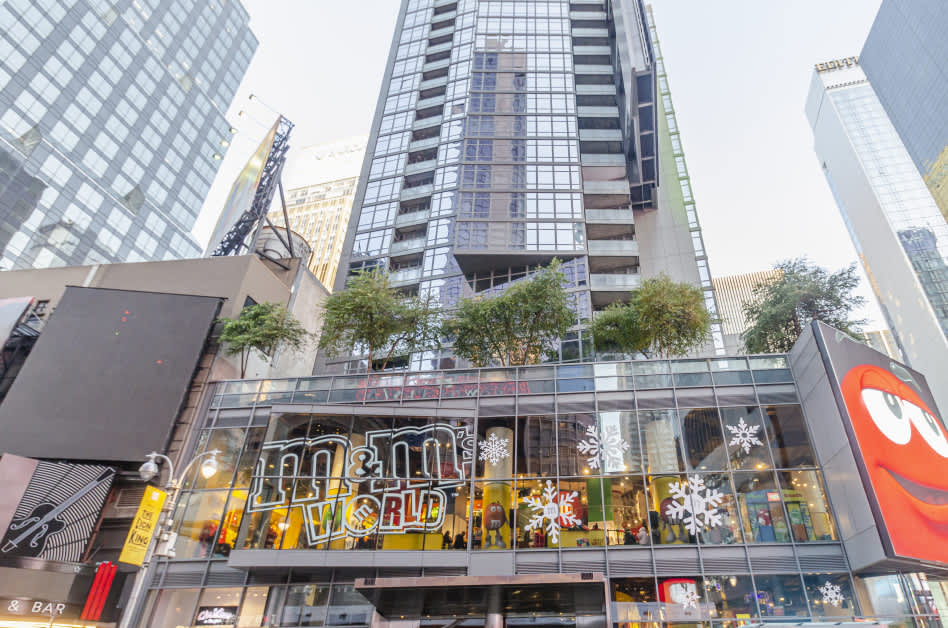 Tall residential tower with glass and metal facade, street-level storefronts including M&M's World, surrounded by other downtown Manhattan buildings.