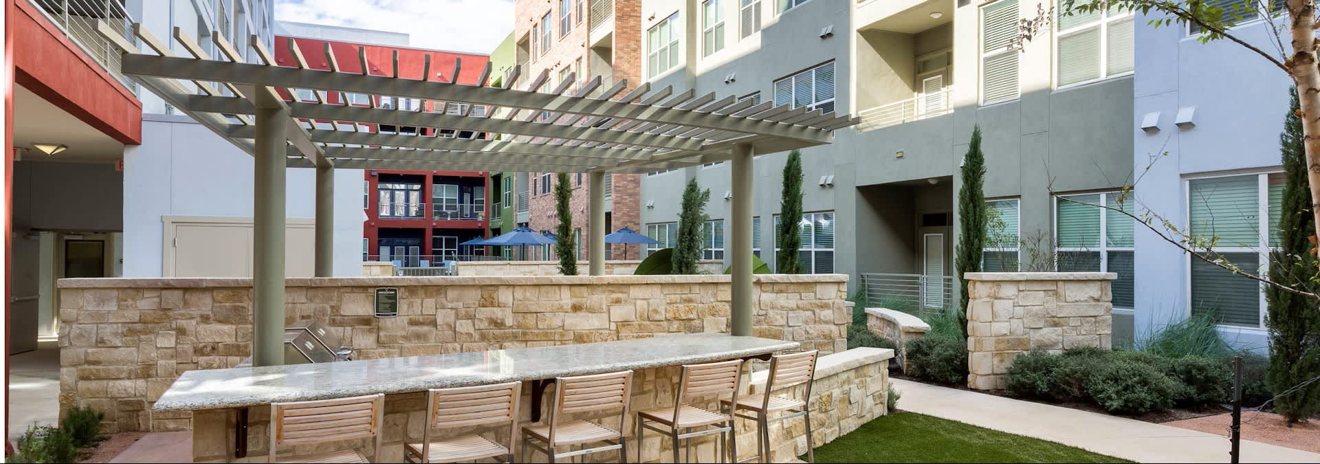 Outdoor courtyard with stone-topped seating counter, wooden chairs, pergola overhead, and residential buildings surrounding green space.