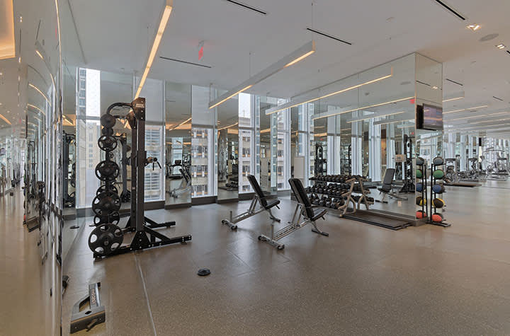 Modern gym with weight racks, exercise bikes, and machines; floor-to-ceiling windows overlook city skyline; bright linear ceiling lights.