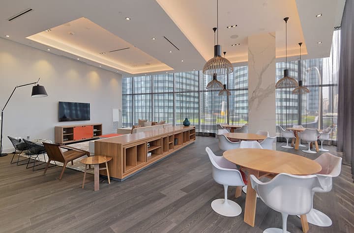 Modern open-concept lounge with wood dining tables, white chairs, pendant lighting, wall-mounted TV, and floor-to-ceiling windows overlooking downtown Toronto.