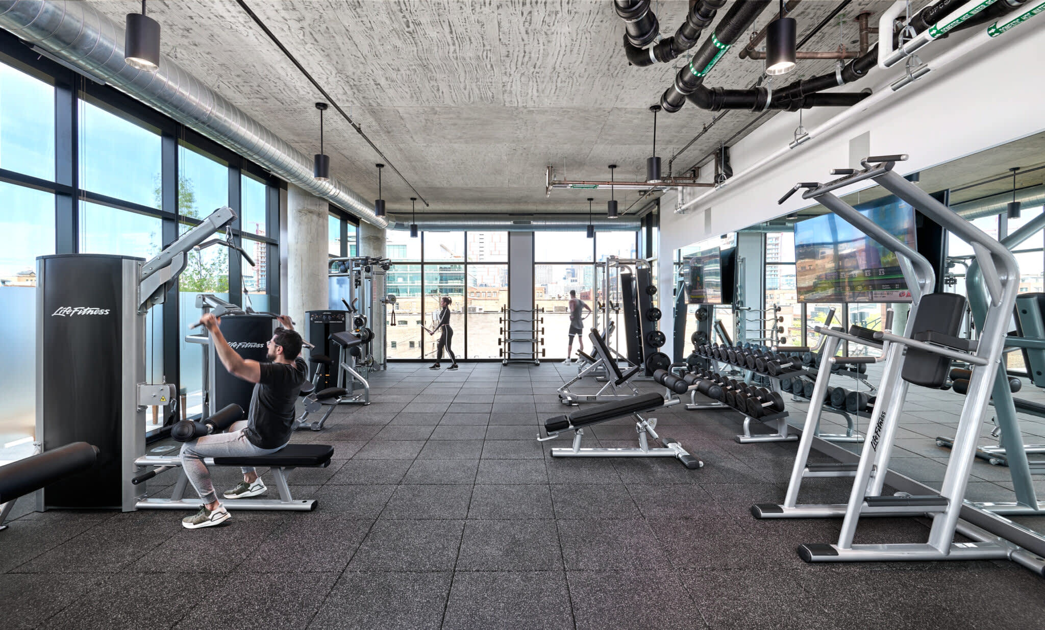 Fitness center with cardio and strength-training equipment, large windows overlooking the city, concrete ceiling with exposed ductwork.