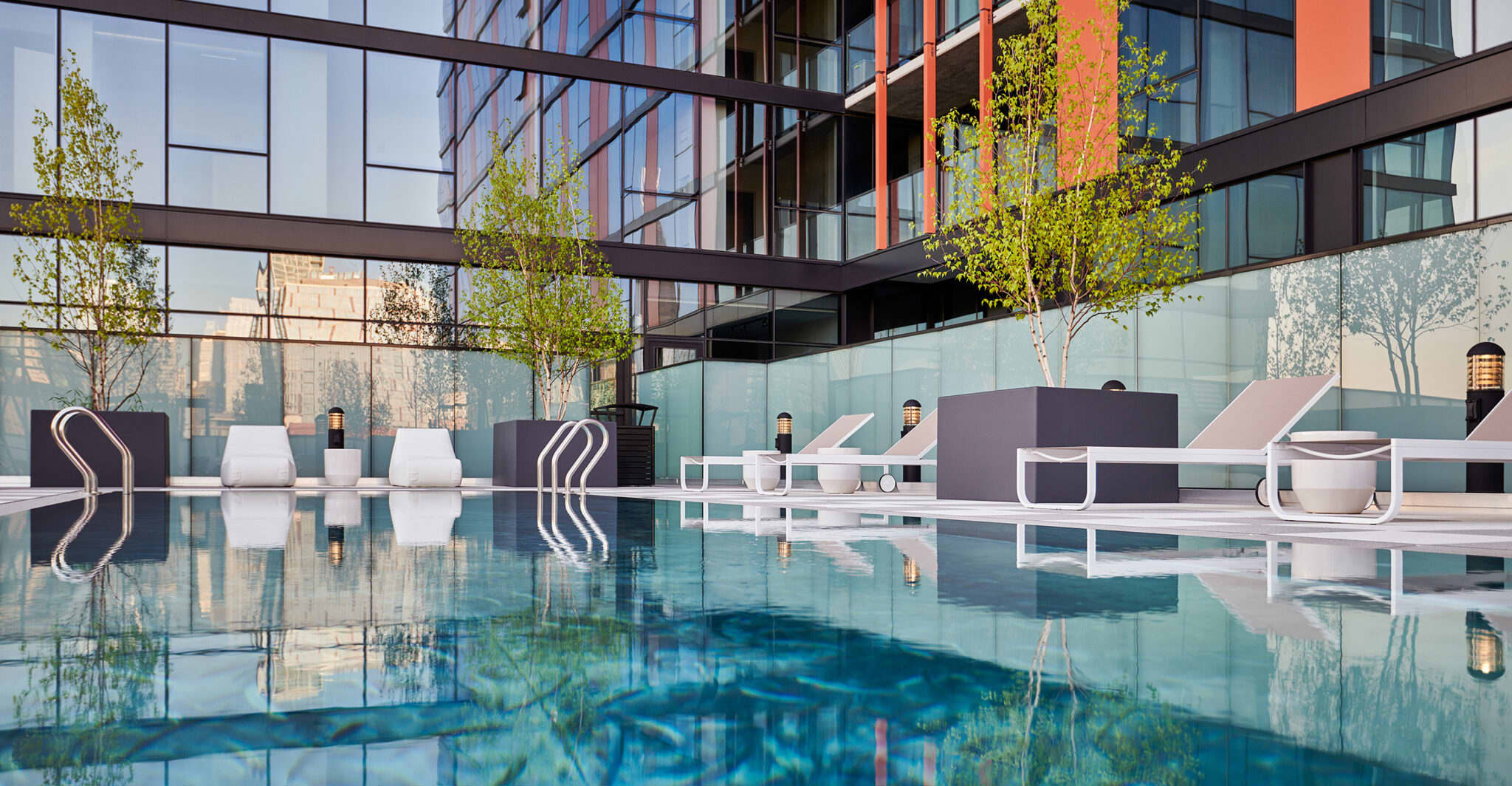 Rooftop pool with clear water, white lounge chairs, dark privacy screens, potted trees, and modern high-rise building backdrop.