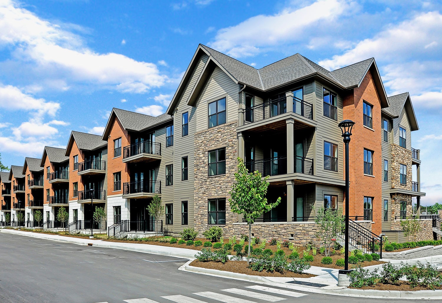 Multi-story residential building with brick and tan siding, black balconies, stone accents, and manicured landscaping along street.