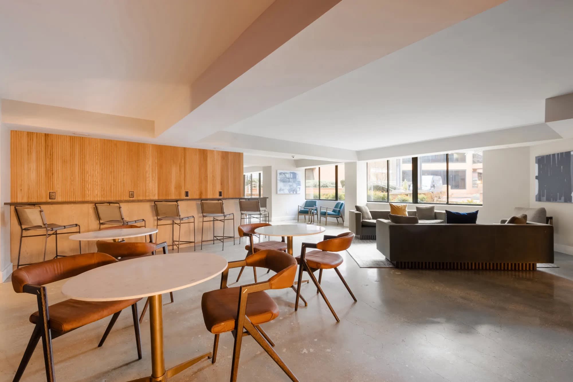 Open-concept common room with mid-century modern furniture, wood-paneled wall, round tables with orange chairs, and large windows overlooking the city.