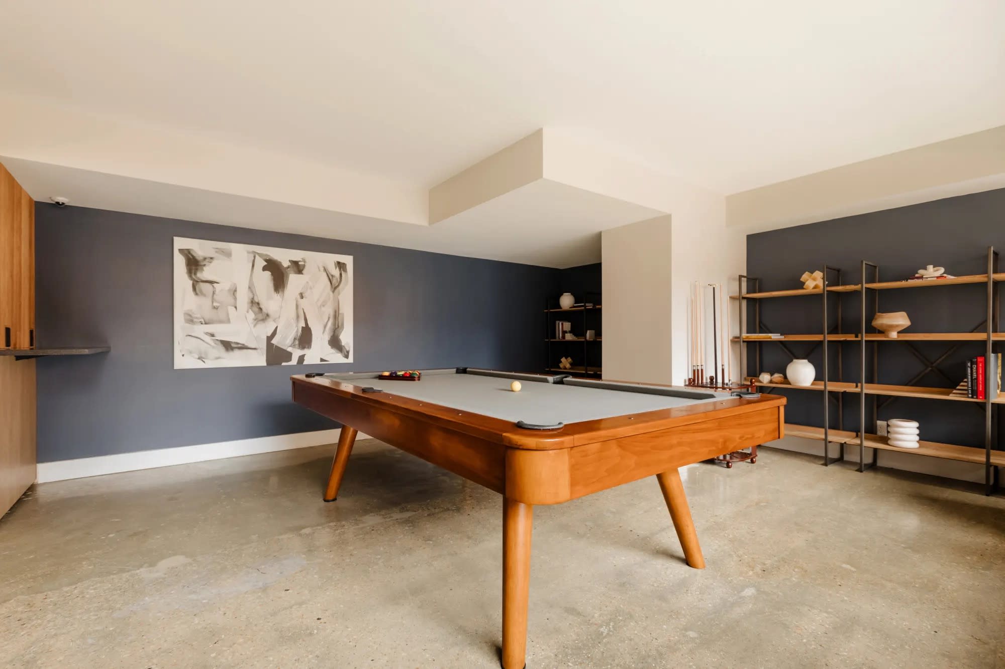 Recreation room with mid-century wood pool table, dark blue accent wall, floating wooden shelves, and framed artwork.