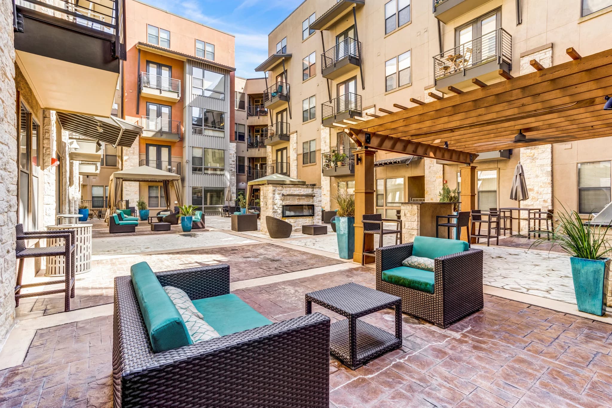 Courtyard with woven lounge chairs, turquoise cushions, wooden pergola, fireplace, and multi-story residential buildings under blue sky.