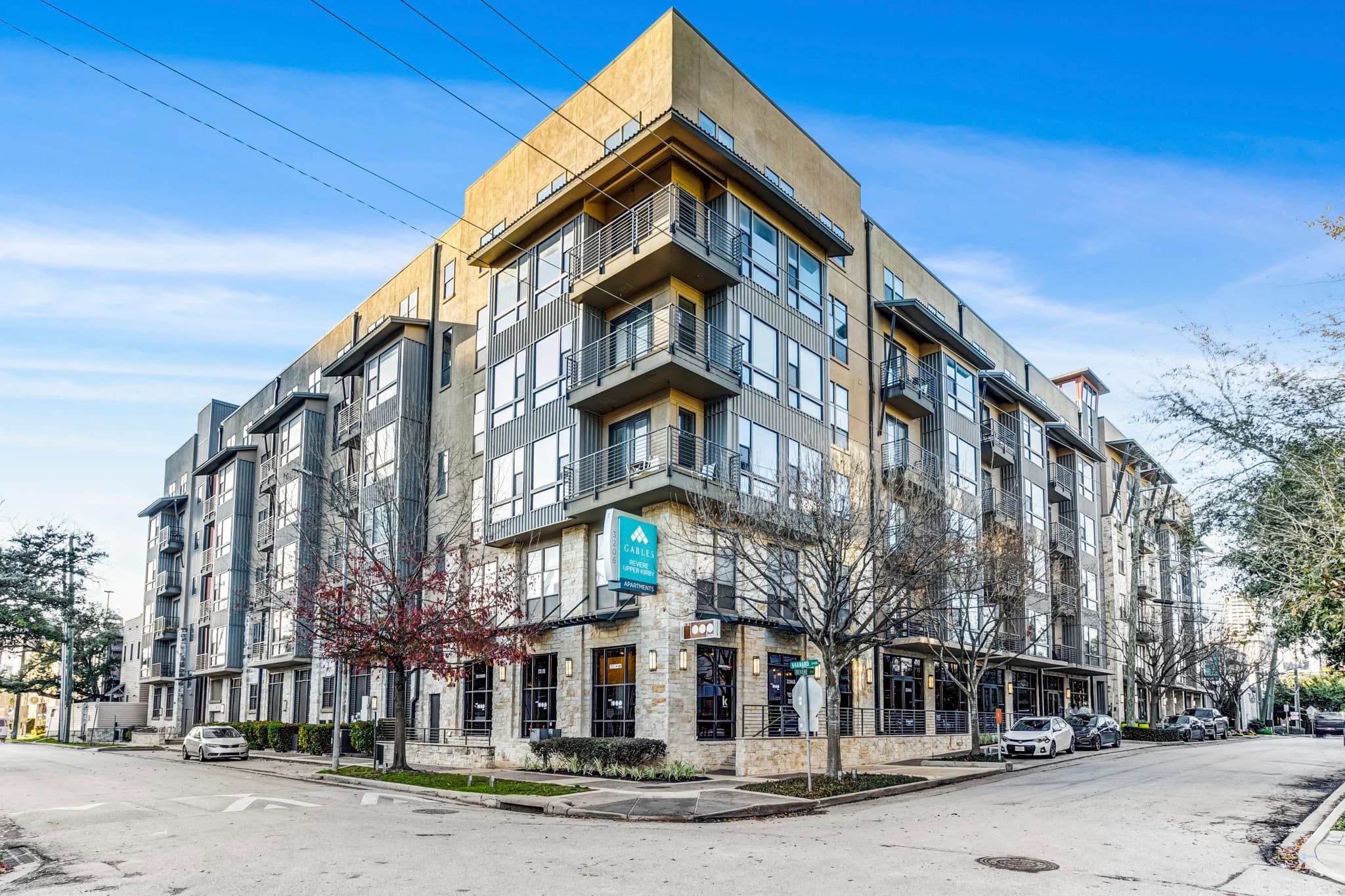 Corner view of a contemporary multi-story residential building with tan and gray facade, balconies, ground-floor retail storefronts, and street-level parking.
