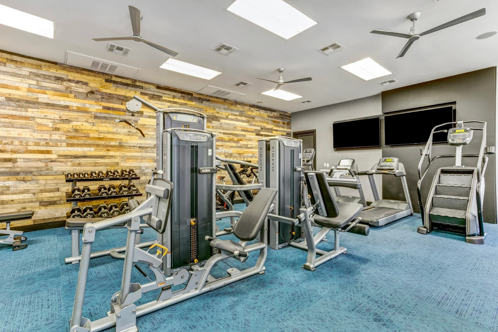 Fitness center with cardio machines, weight racks, and strength training equipment on blue flooring beneath skylights and wood-paneled walls.