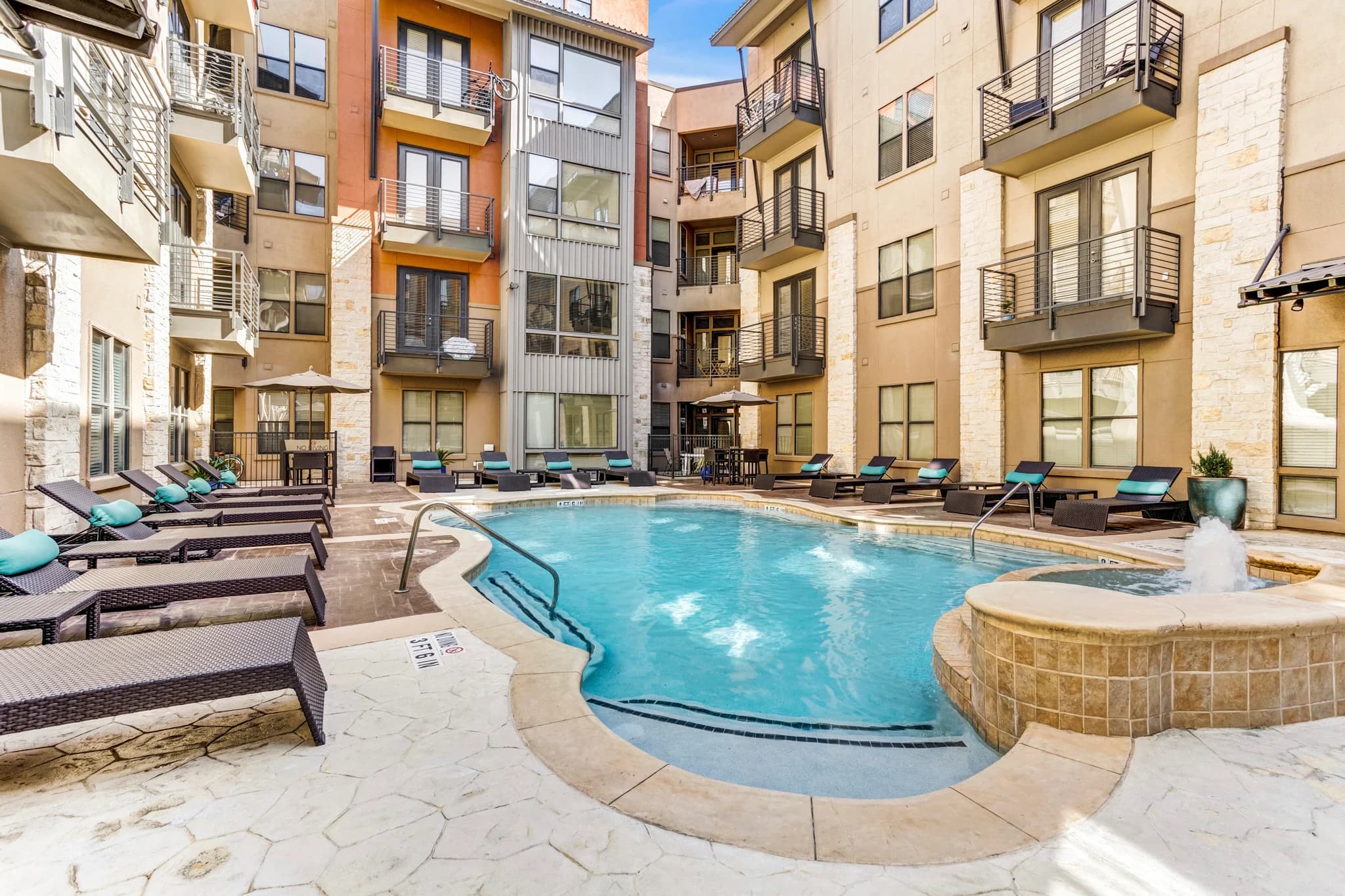 Resort-style swimming pool with attached spa, lounge chairs, and multi-story apartment buildings with balconies in background.