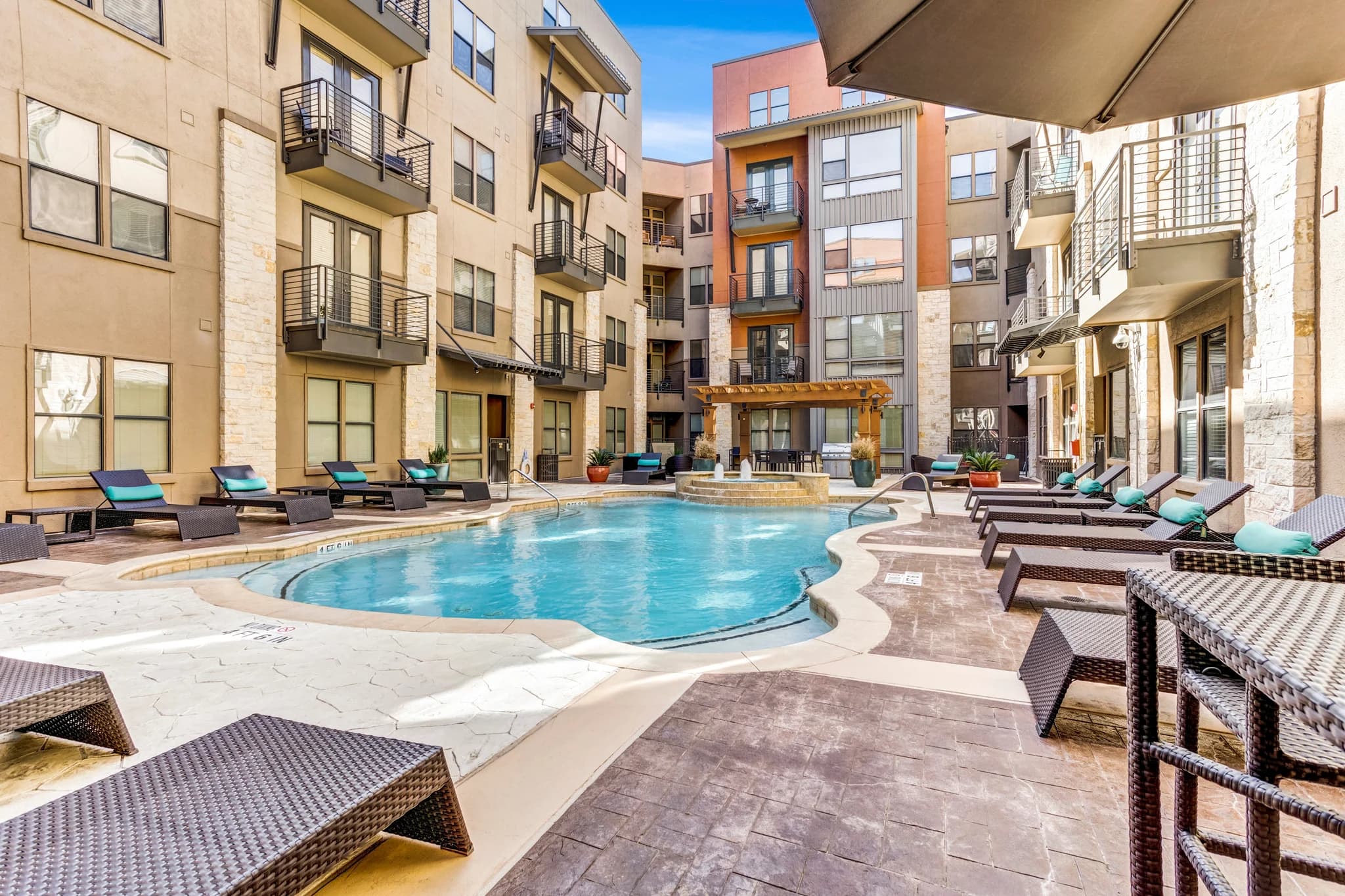 Curved swimming pool with turquoise water, surrounded by lounge chairs, patio seating, and multi-story residential buildings under clear blue sky.