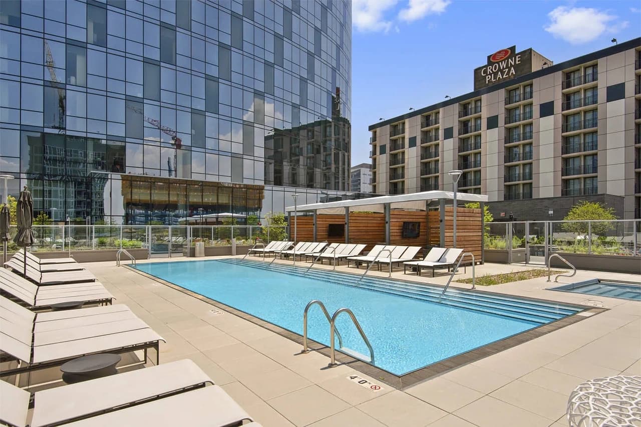 Outdoor rooftop pool with lounge chairs, wooden cabanas, and surrounding high-rise buildings under blue sky.