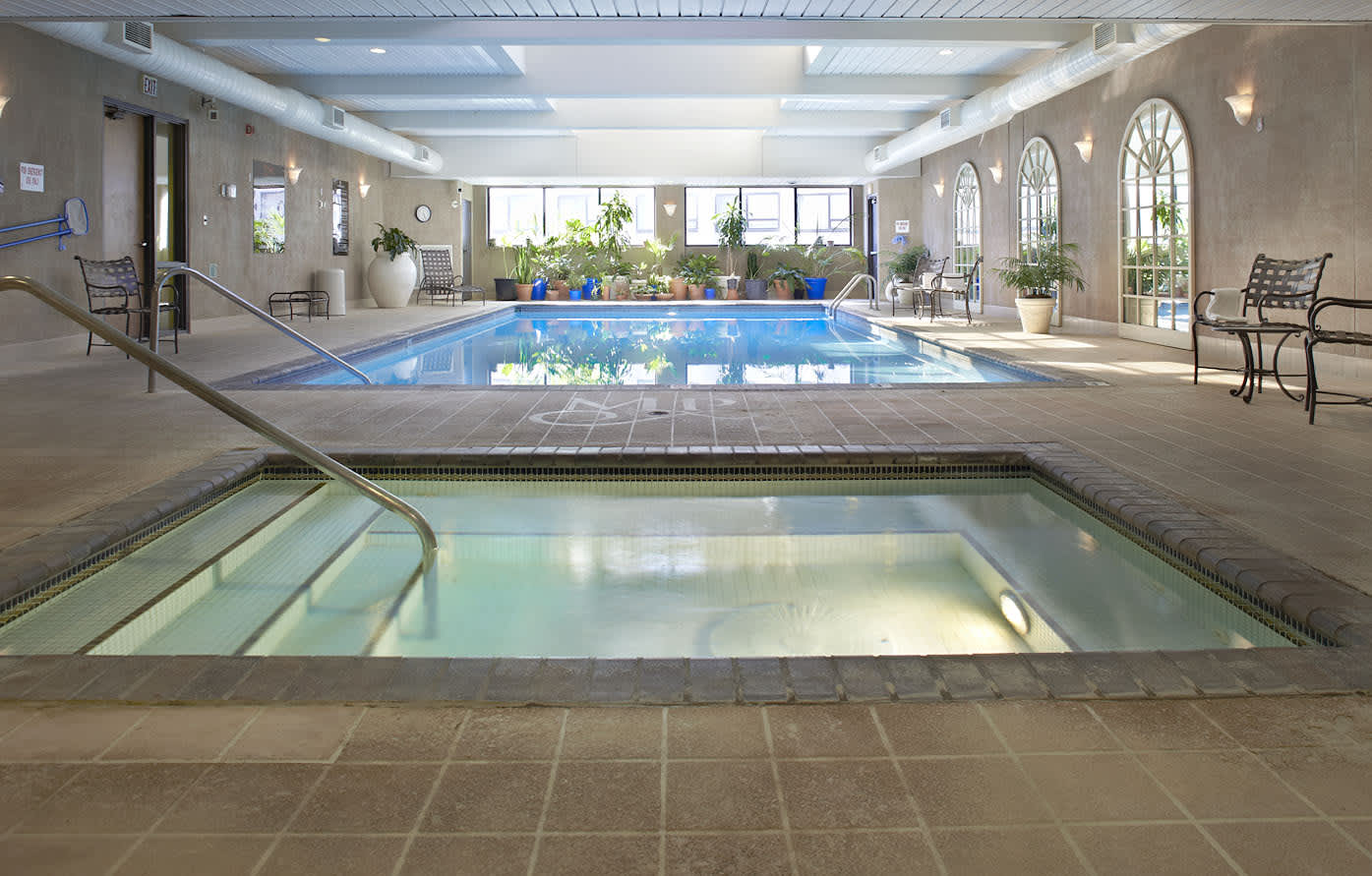 Indoor pool facility with two pools, one in foreground with handrails and one in background, surrounded by lounge chairs, potted plants, and large windows.