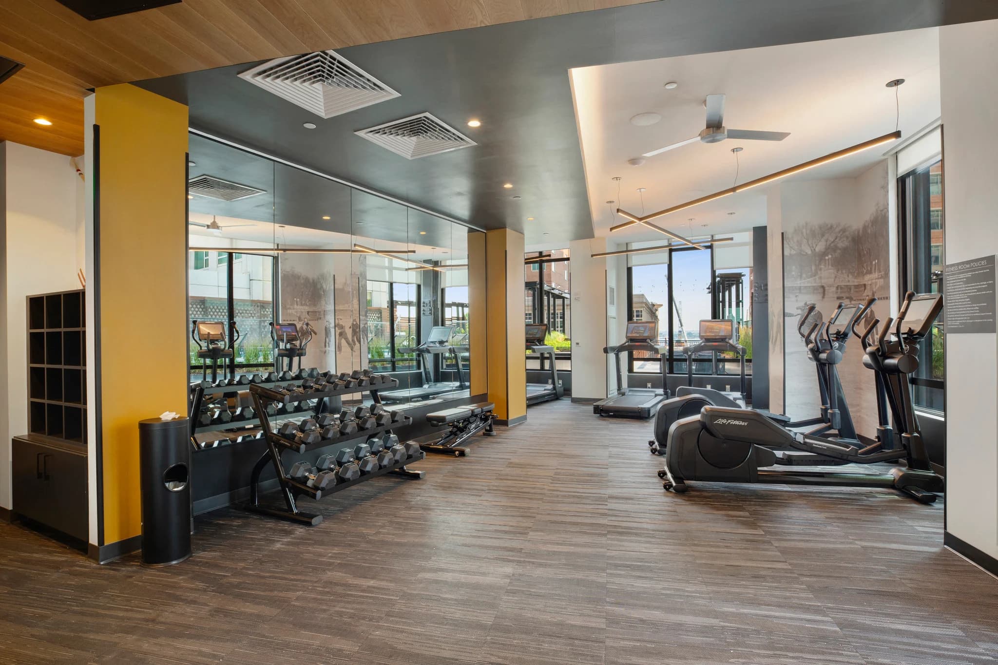 Fitness center with dumbbell racks, elliptical machines, and large windows with city views, wood flooring, yellow accent column, and gray ceiling.
