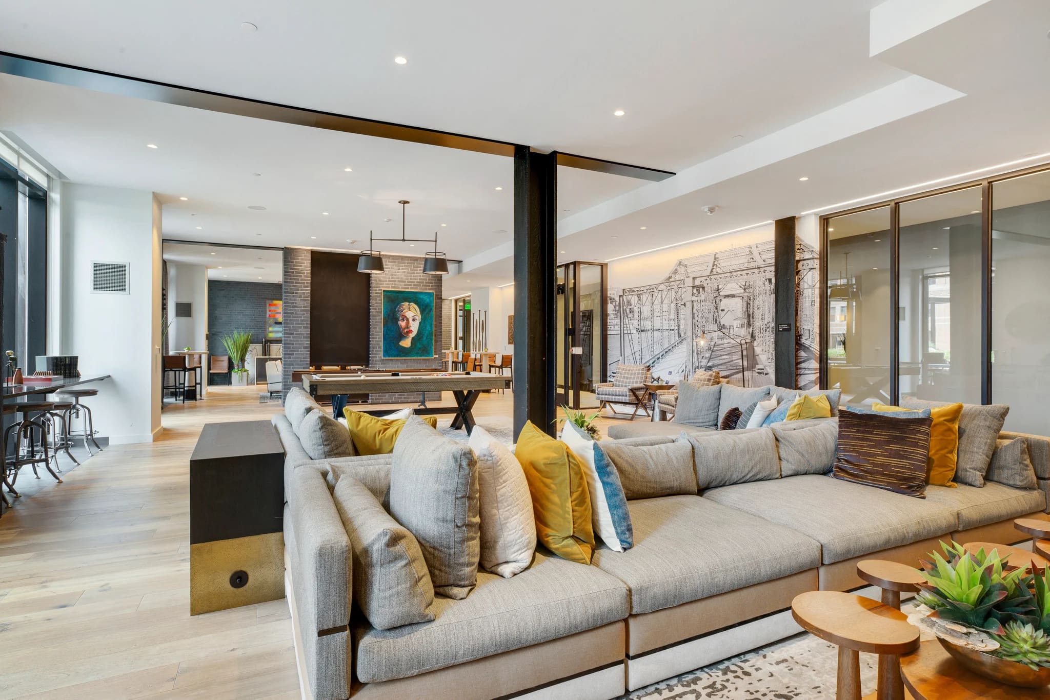 Open living room with gray sectional sofa, yellow accent pillows, light wood flooring, and exposed brick walls with modern artwork.